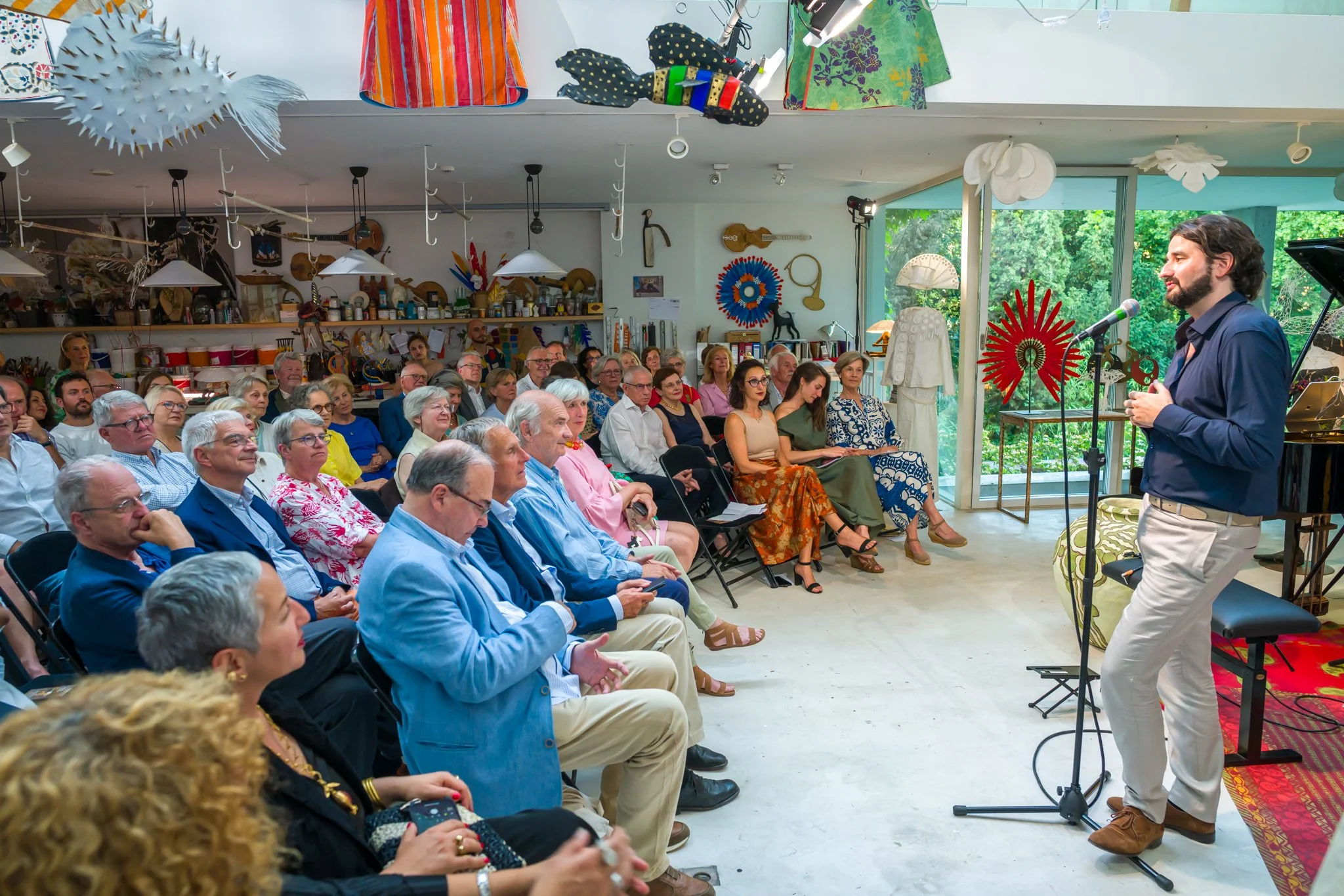 Un homme parle devant un public dans une pièce décorée d'objets d'art et d'instruments de musique, avec un grand vase rouge derrière lui.