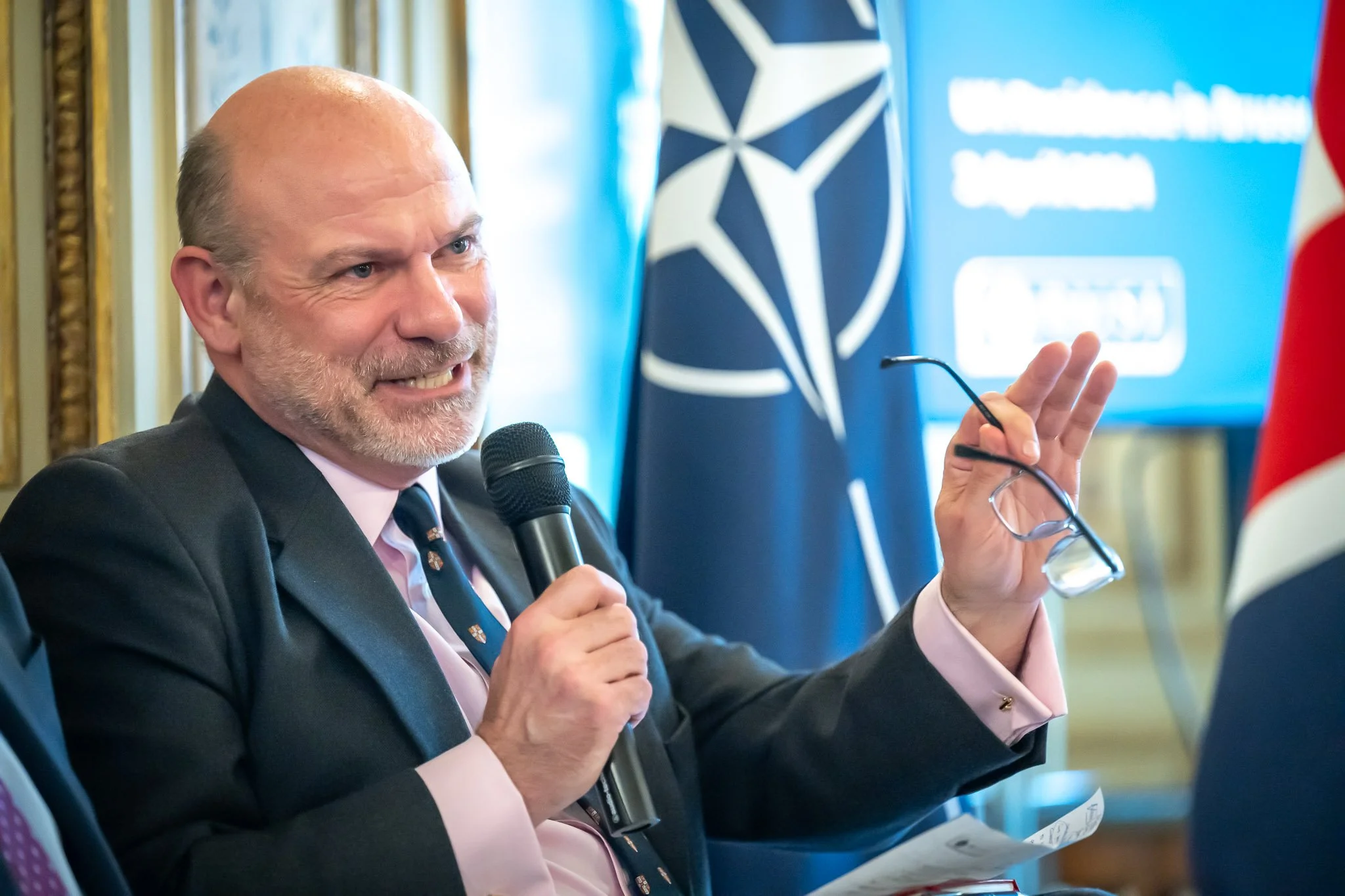 A man holding a microphone and a pair of glasses, with a NATO flag in the background.