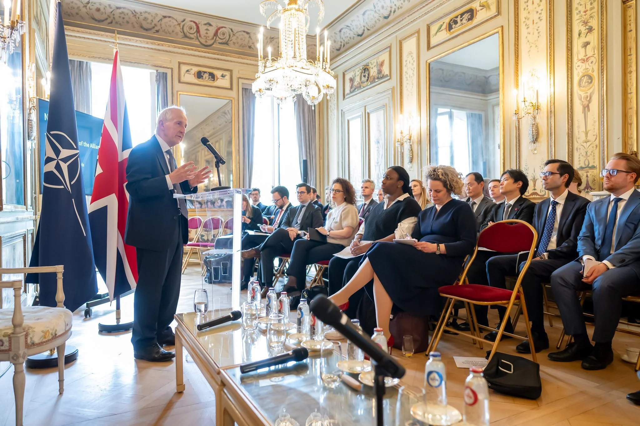 A man makes a speech to a seated audience in an elegant room with mirrors and chandeliers. NATO and UK flags are visible behind him.