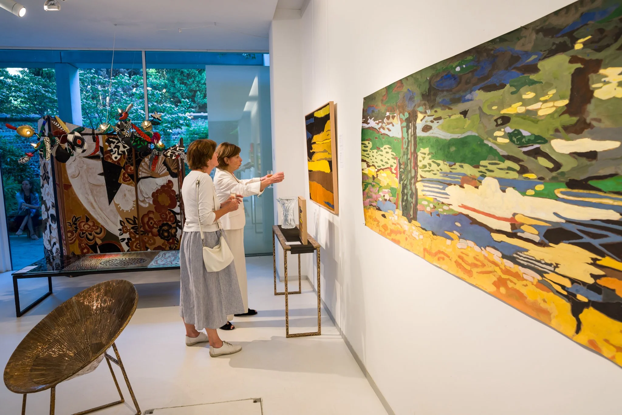 Deux femmes regardent et discutent de peintures dans une galerie d'art moderne, avec une œuvre impressionniste aux couleurs vives accrochée au mur à leur droite.