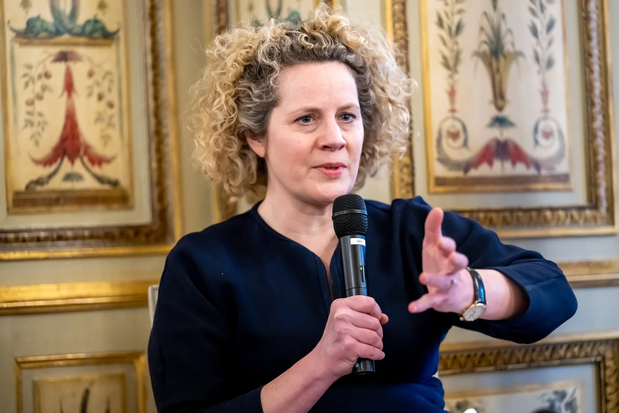 A person holding a microphone, speaking in a room decorated with elaborate murals.
