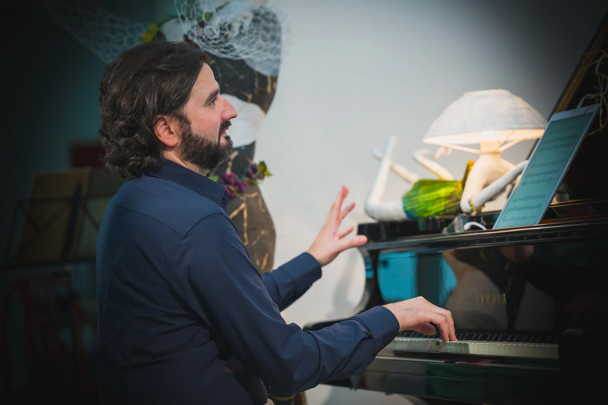 Un homme joue du piano noir Yamaha en regardant une tablette, avec une sculpture de lampadaire et un lampadaire lumineux en fond.