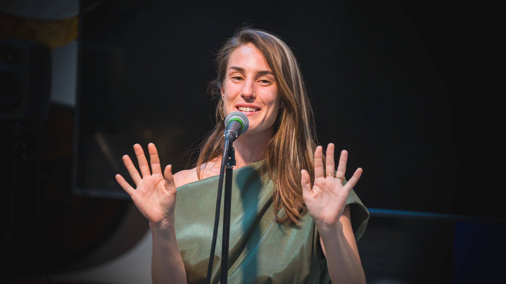 Femme souriante parlant dans un microphone, mains levées, portant une robe verte, fond sombre.