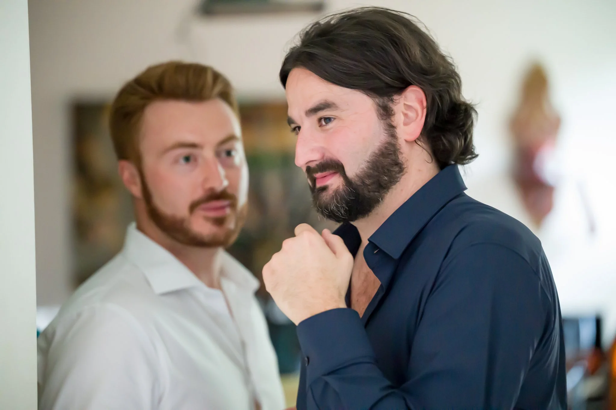 Deux hommes en conversation dans une pièce bien éclairée, l'un porte une chemise blanche et l'autre une chemise bleue foncée, tous deux avec des barbes et des cheveux bruns, dans un environnement intérieur.