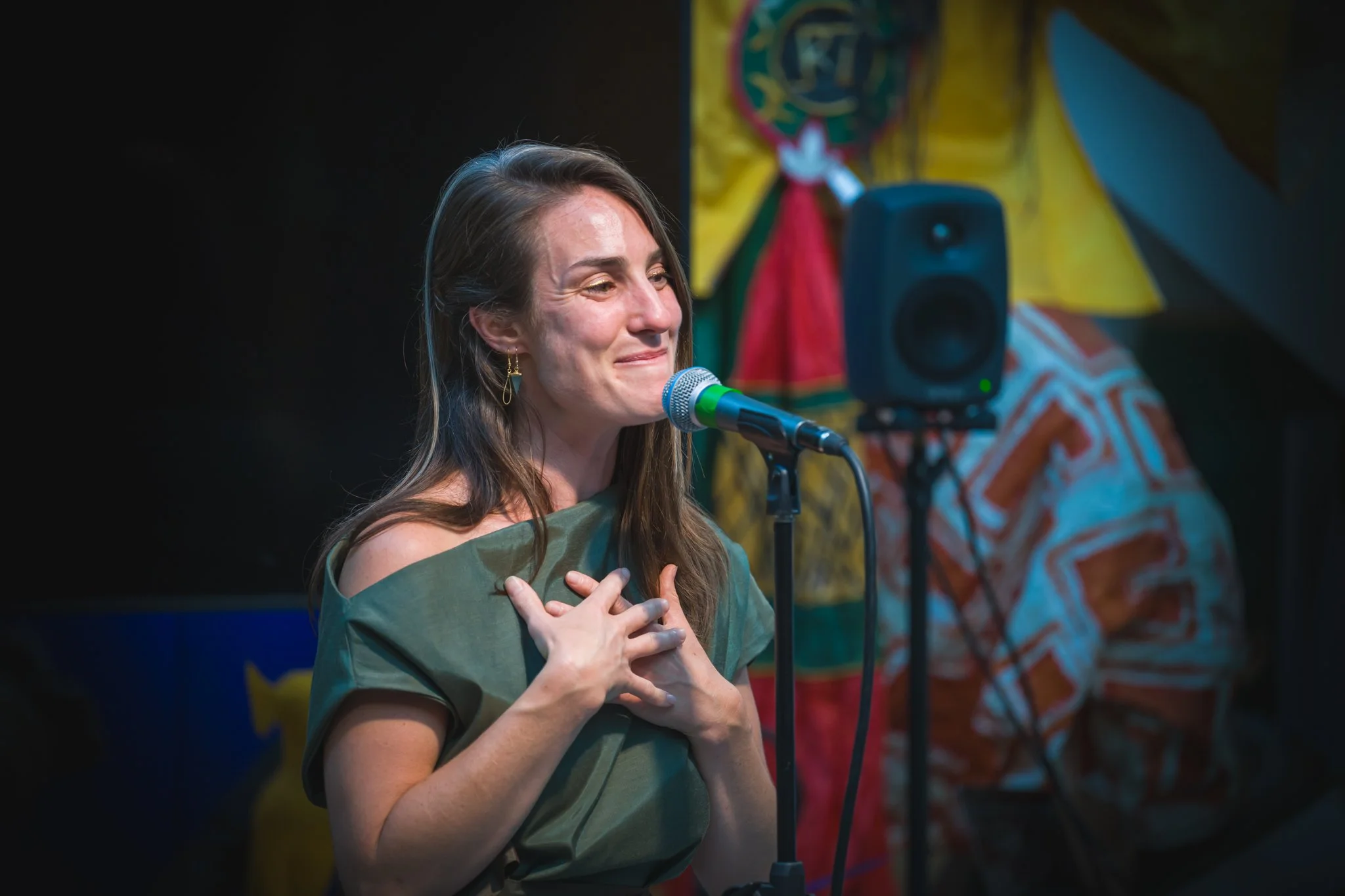 Jeune femme avec longue chevelure brune, en pull vert, lors d'une performance vocale, main sur la poitrine, devant un microphone sur scène, avec un fond coloré.