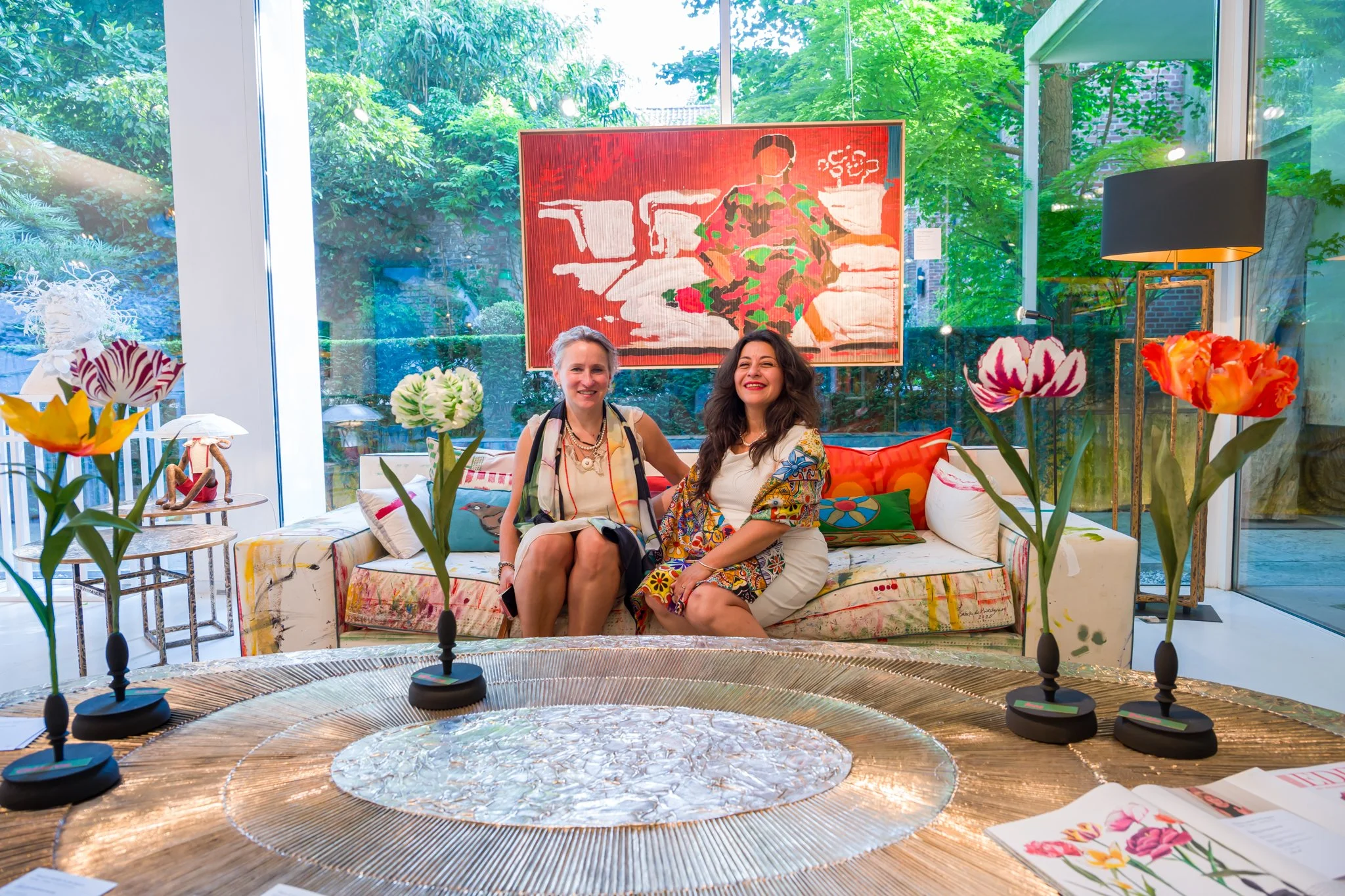 Deux femmes assises sur un canapé dans une pièce lumineuse avec de grandes fenêtres, décorée avec des fleurs en céramique, un tableau en arrière-plan, et des livres posés sur une table en avant.