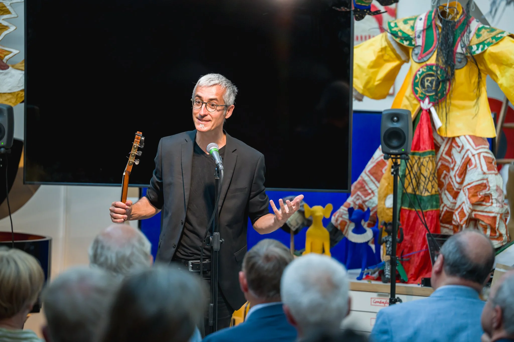 Un homme en costume noir parle avec un micro, tenant une guitare, lors d'une présentation ou performance devant un public. Il y a des figures colorées en arrière-plan et un grand écran noir derrière lui.