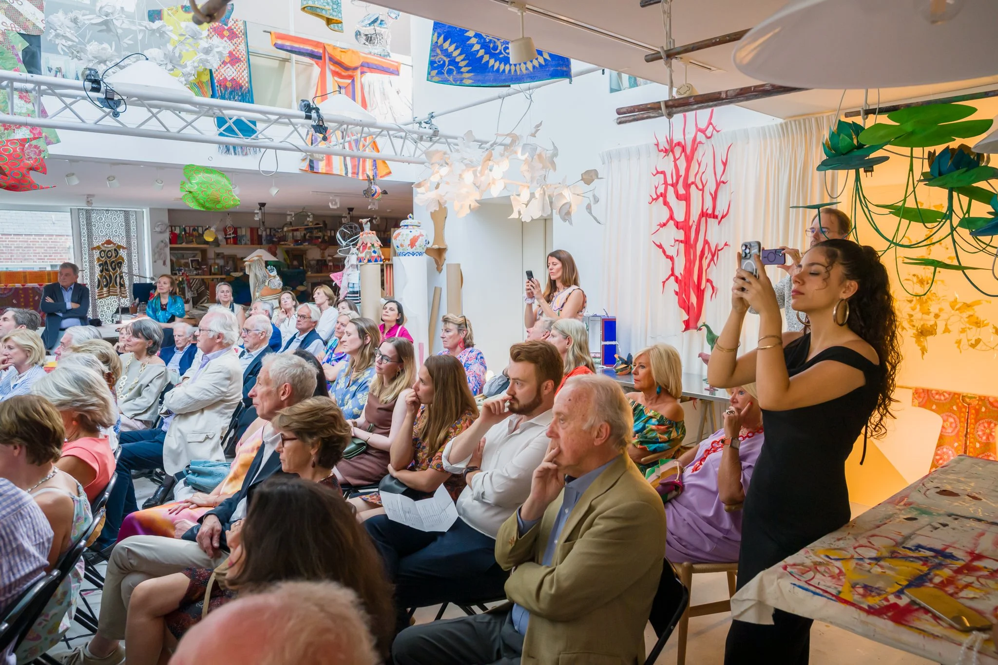 Une salle d'événement avec un public attentif, des femmes prenant des photos avec leur téléphone, des décorations colorées et des œuvres d'art suspendues au plafond.