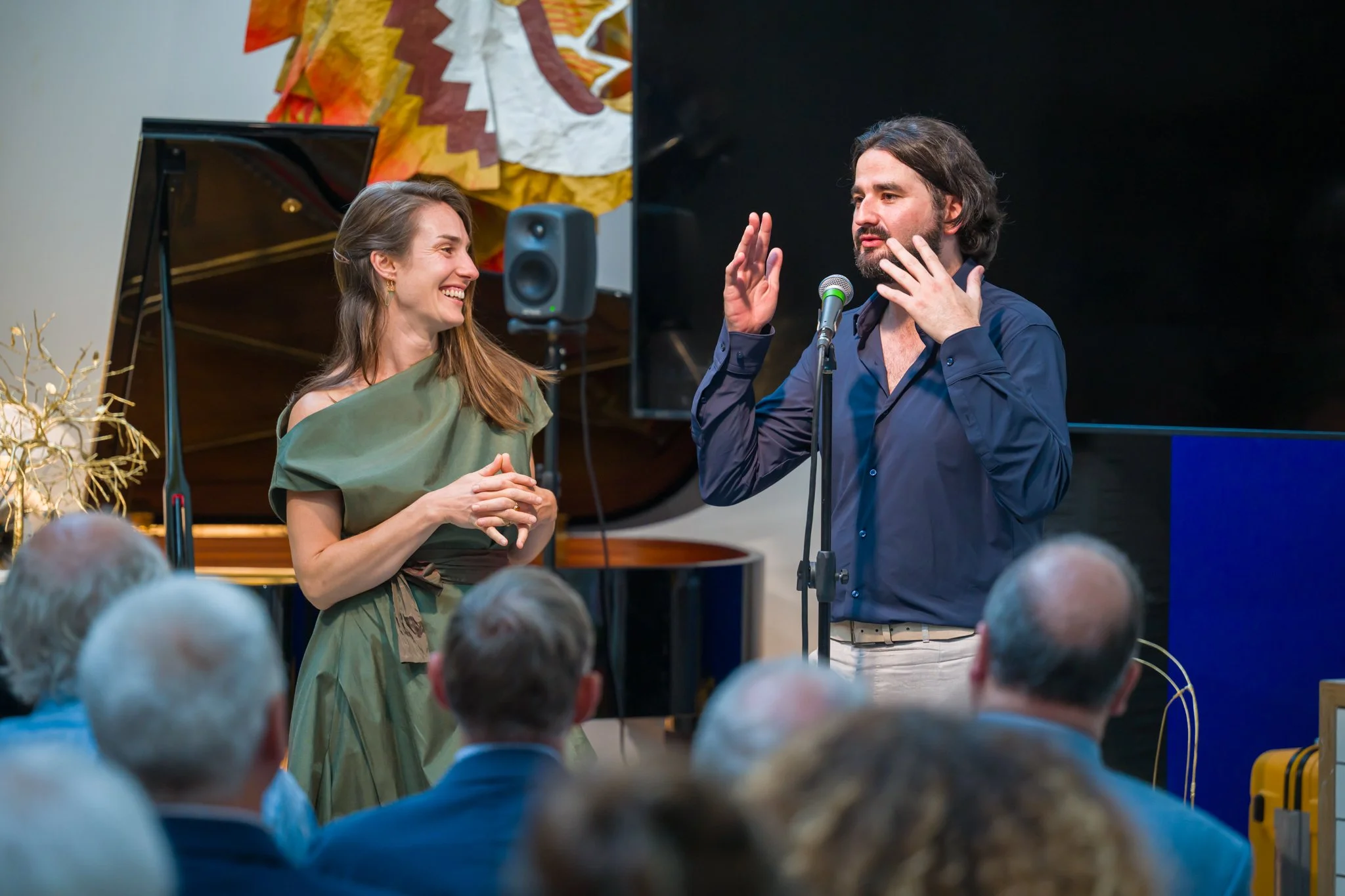 Un homme et une femme parlent lors d'une performance ou d'une conférence, entourés d'un public, avec un piano noir en arrière-plan.