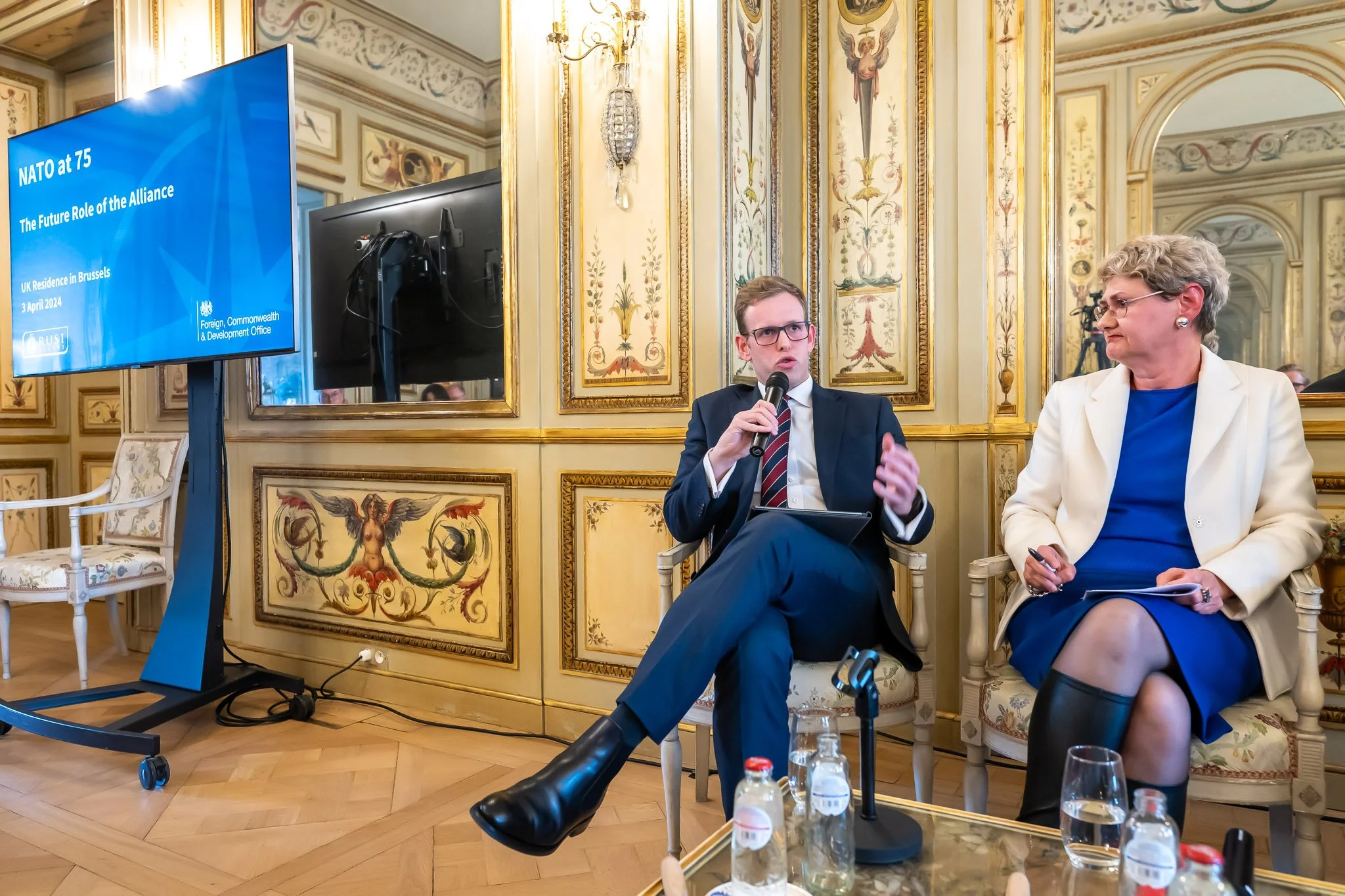 Two people in conversation, vintage interior fashion, elaborate décor, sign reading "NATO at 75: The Future Role of the Alliance".