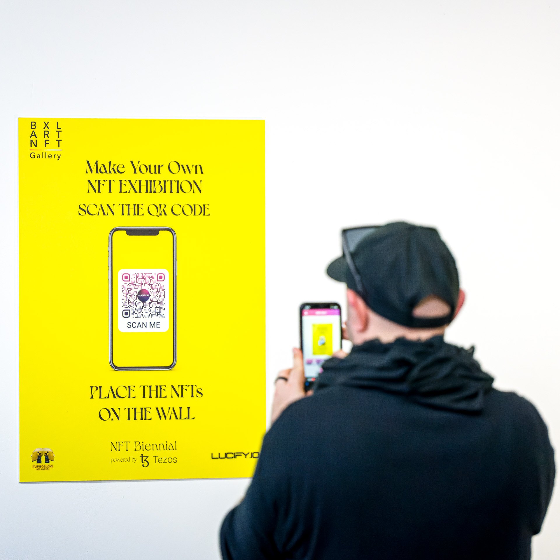 Un homme scanne un code QR sur une affiche jaune qui promeut une exposition NFT à la "Bxlt NFT Gallery". Le poster invite à créer sa propre exposition NFT.