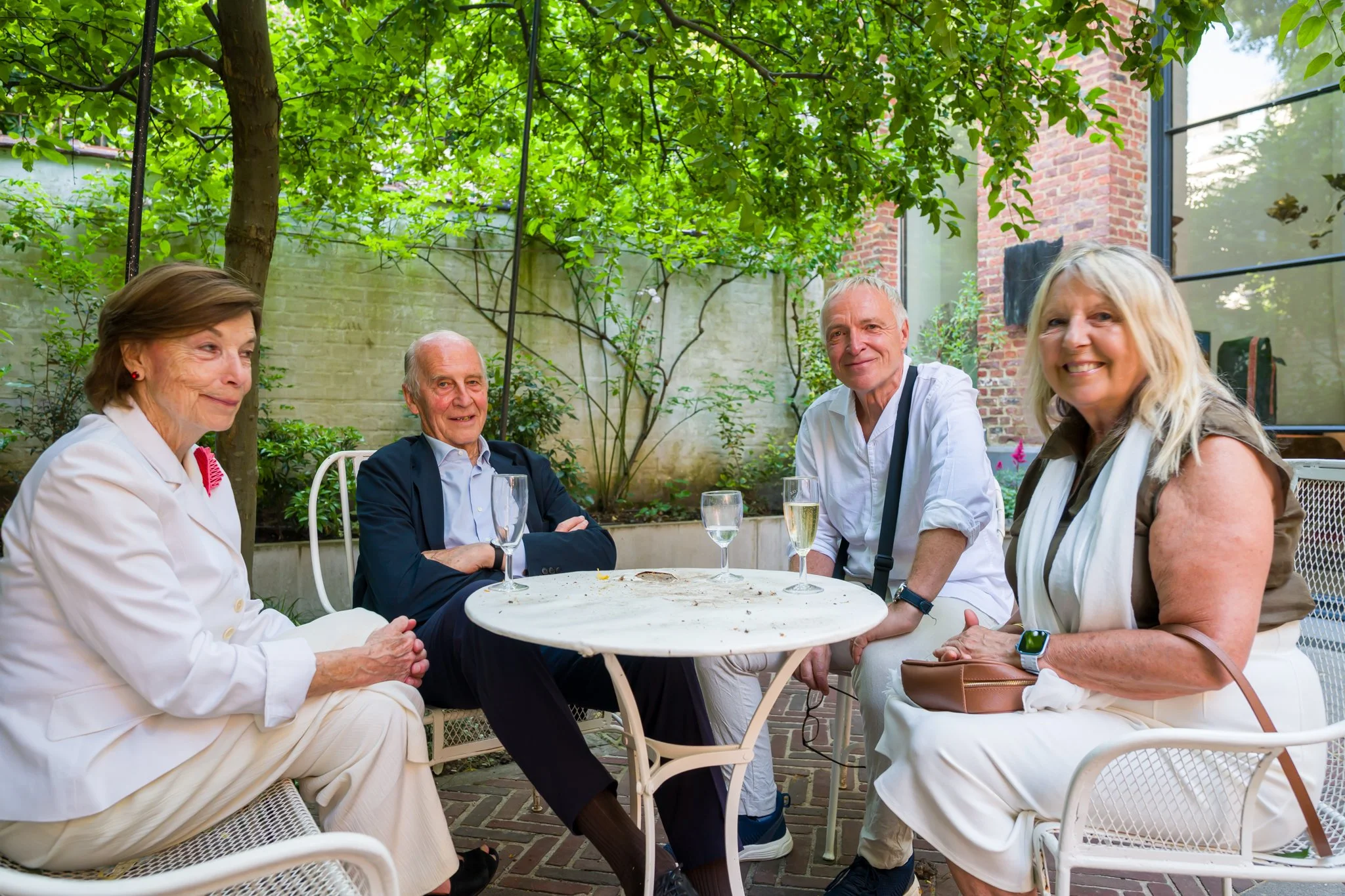 Cinq personnes âgées, trois femmes et deux hommes, assises autour d'une table en terrasse, à l'extérieur, avec des verres de champagne ou de vin blanc, sous un arbre vert, dans un environnement de jardin ou patio.