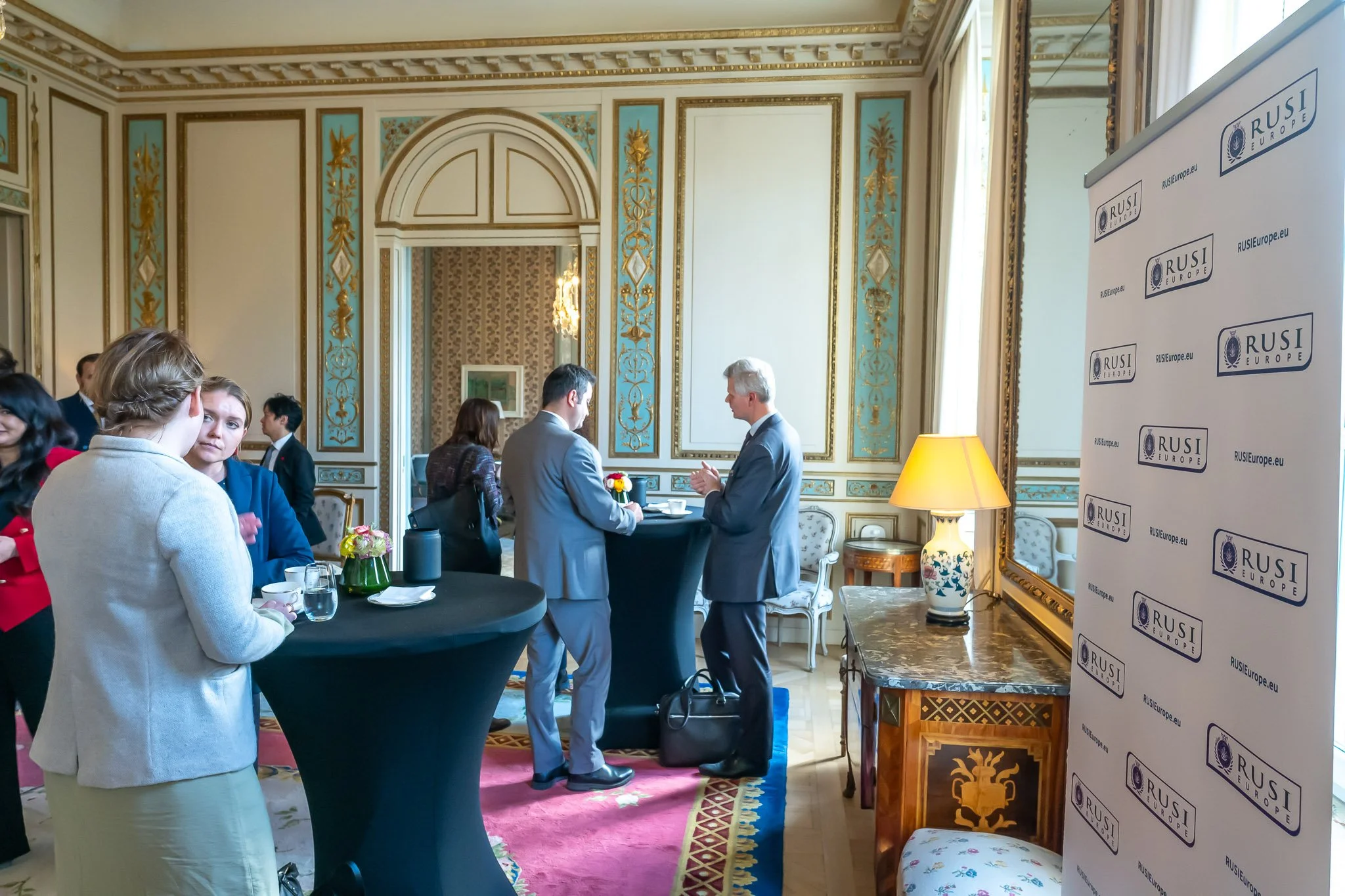 A group of people stand and chat in an ornate room at a networking event, with a "RUSI Europe" banner visible on the right.