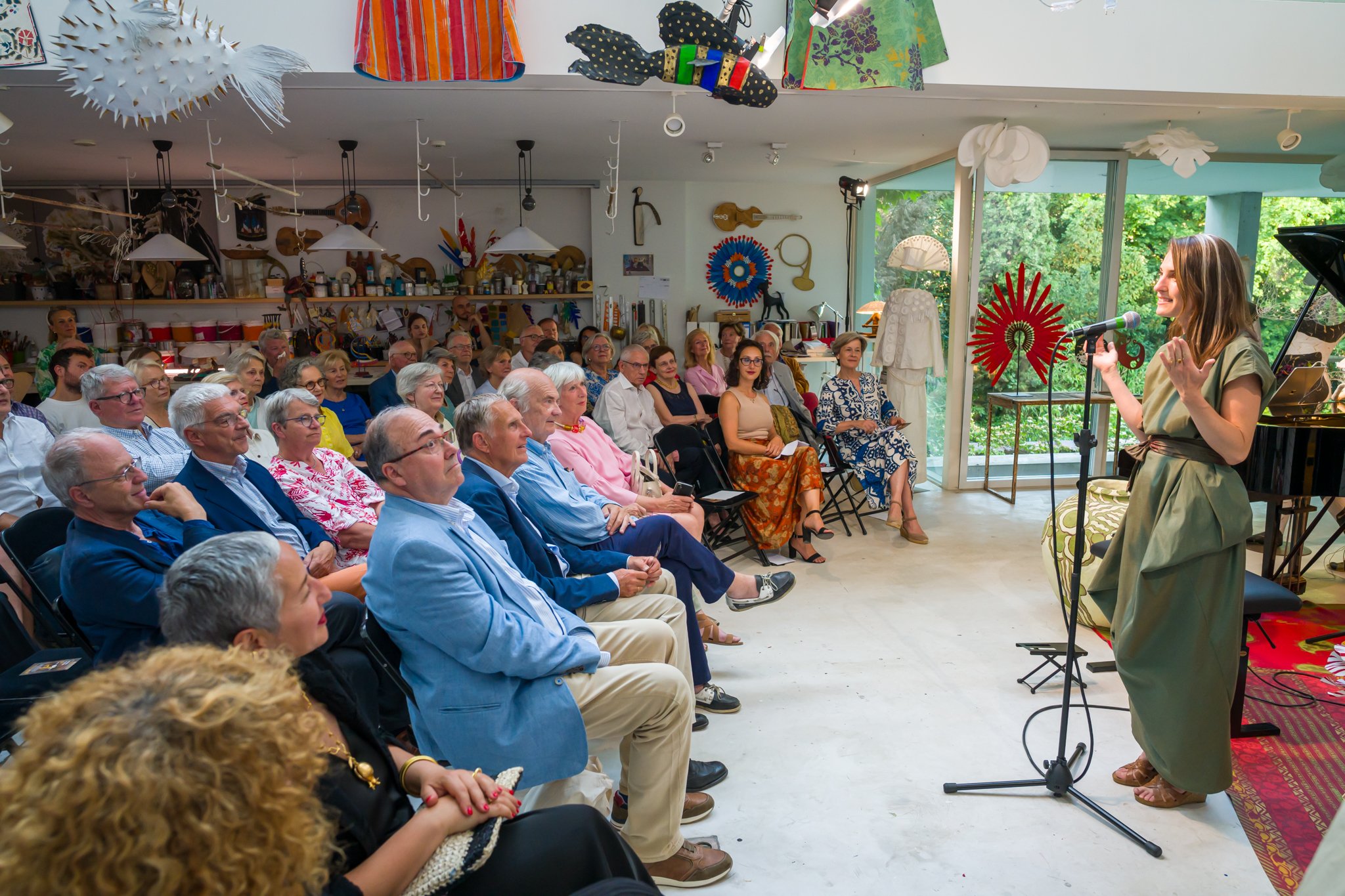 Une femme chante ou parle en public lors d'une performance ou d'une présentation devant un public assis dans une salle décorée avec des objets artistiques et artisanaux. Le public est attentif, et la scène est située près d'une grande fenêtre donnant
