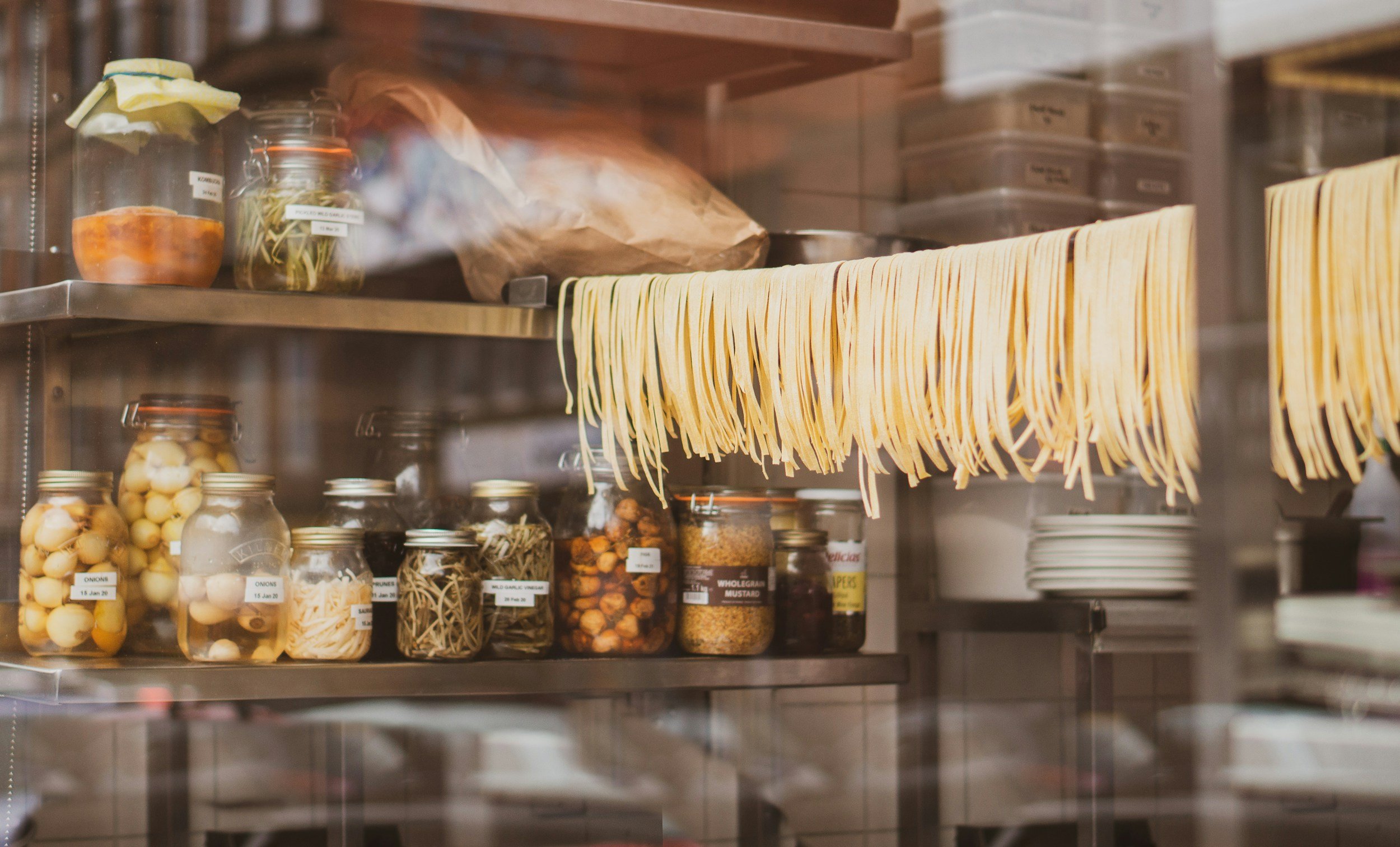 Dry spaghetti hanging on a rack inside a pantry, with jars of preserved vegetables, onions, mustard, and other canned goods on shelves below.
