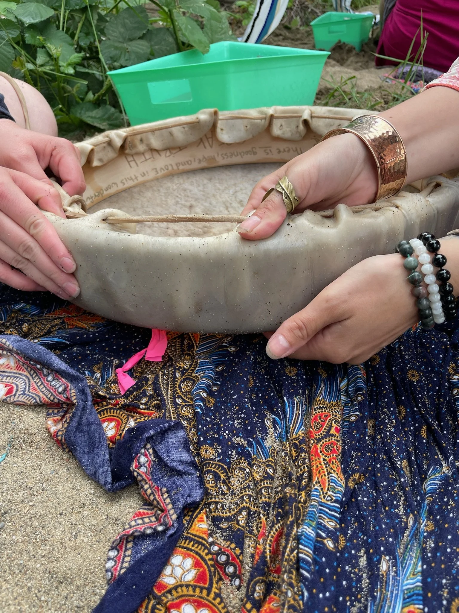 Close-up of two hands holding a large, beige, ceramic bowl or dish outdoors on sand, with green plants and containers in the background. One hand has a gold bracelet and rings, and several bracelets, including black, white, and gray beads. The other hand appears to be guiding or stabilizing the bowl.