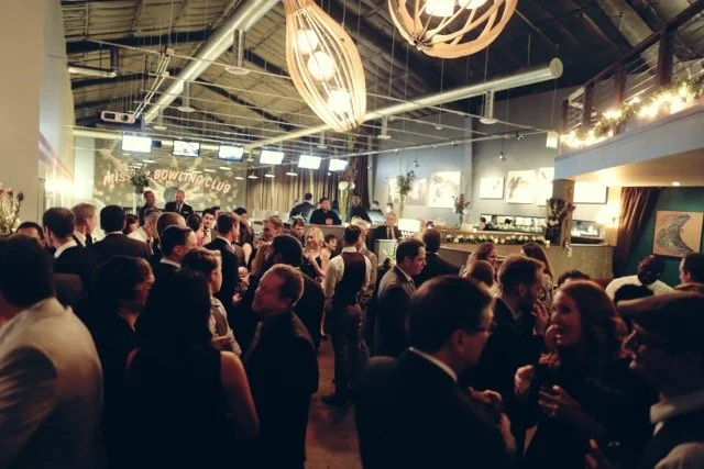 A bride and groom holding hands and dancing at their wedding reception, surrounded by guests who are smiling, taking photos, and clapping in a dimly lit venue with artwork on the walls.