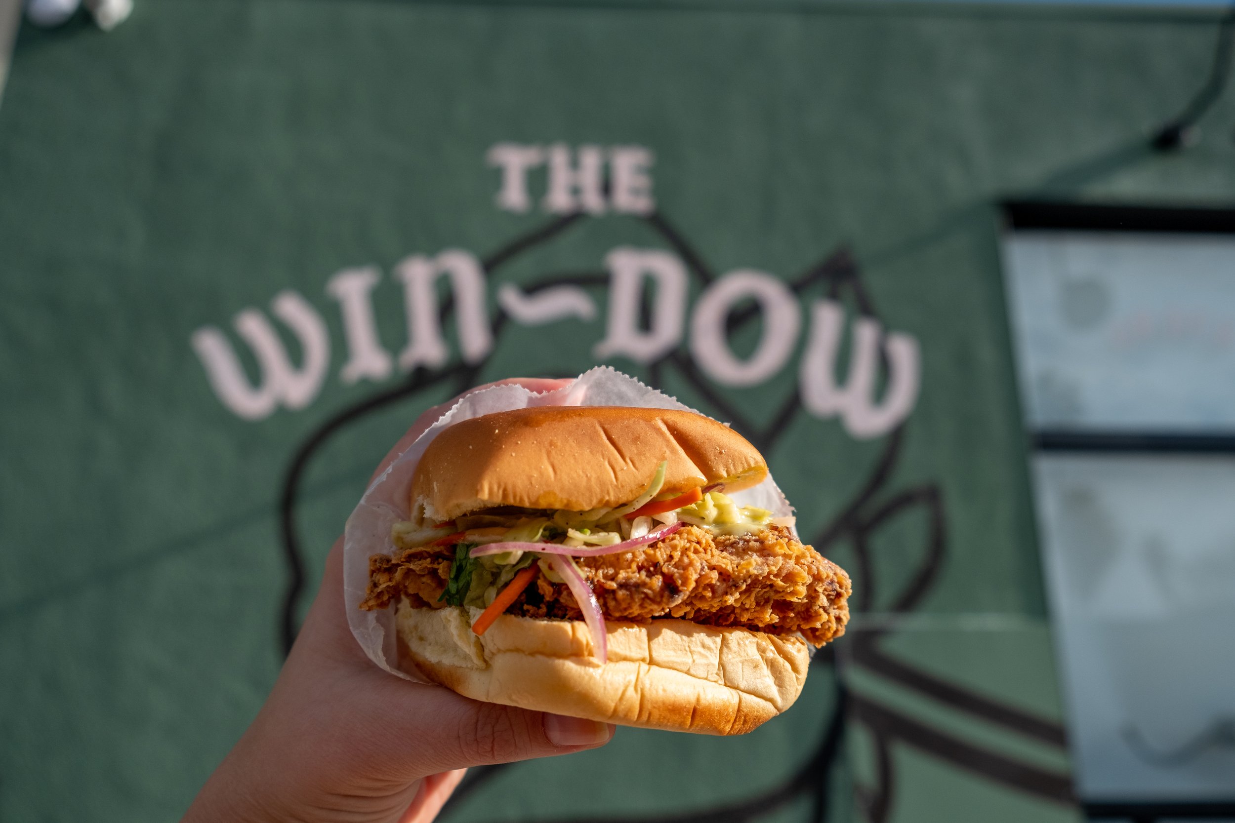  hand holding fried chicken sandwich in front of a mural of the Win~Dow logo 