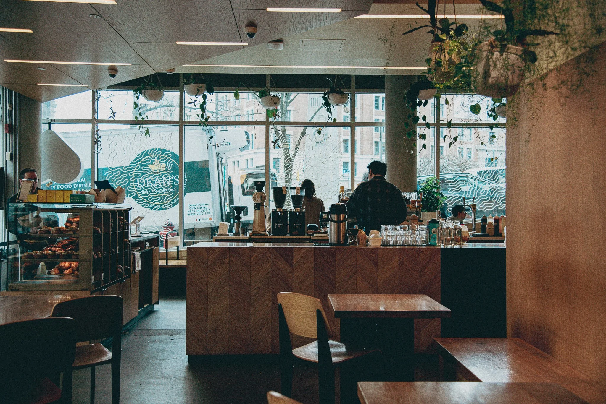 Sitting inside Nemesis coffee in Vancouver, BC. Looking at the barista carefully preparing our morning coffee with a view out the window and a pastry case in the corner.