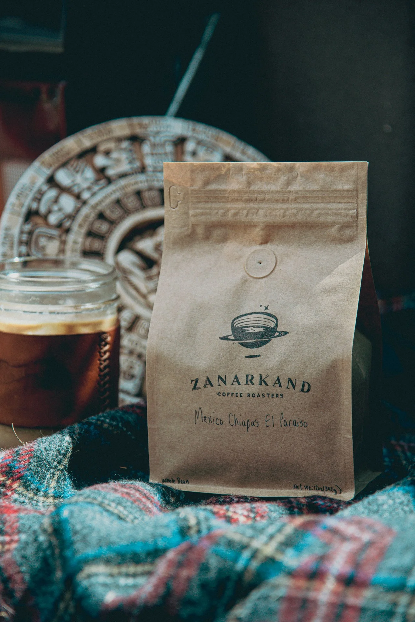 A brown paper coffee bag labeled with 'zanarkand' and an Mexican calendar in the background with a Pendleton blanket and iced coffee.