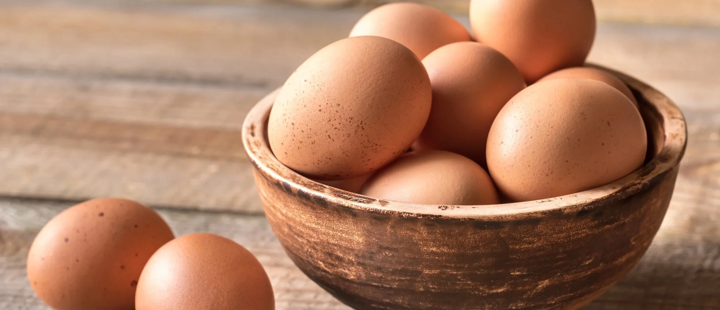 A rustic bowl filled with brown eggs on a wooden surface, with a few eggs outside the bowl.
