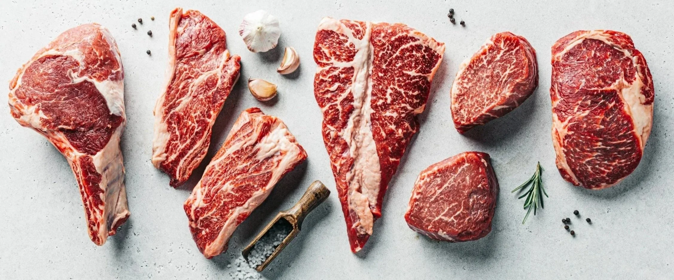 Assorted cuts of raw beef steaks on a light surface, garnished with garlic cloves, black peppercorns, a sprig of rosemary, and a salt shaker.