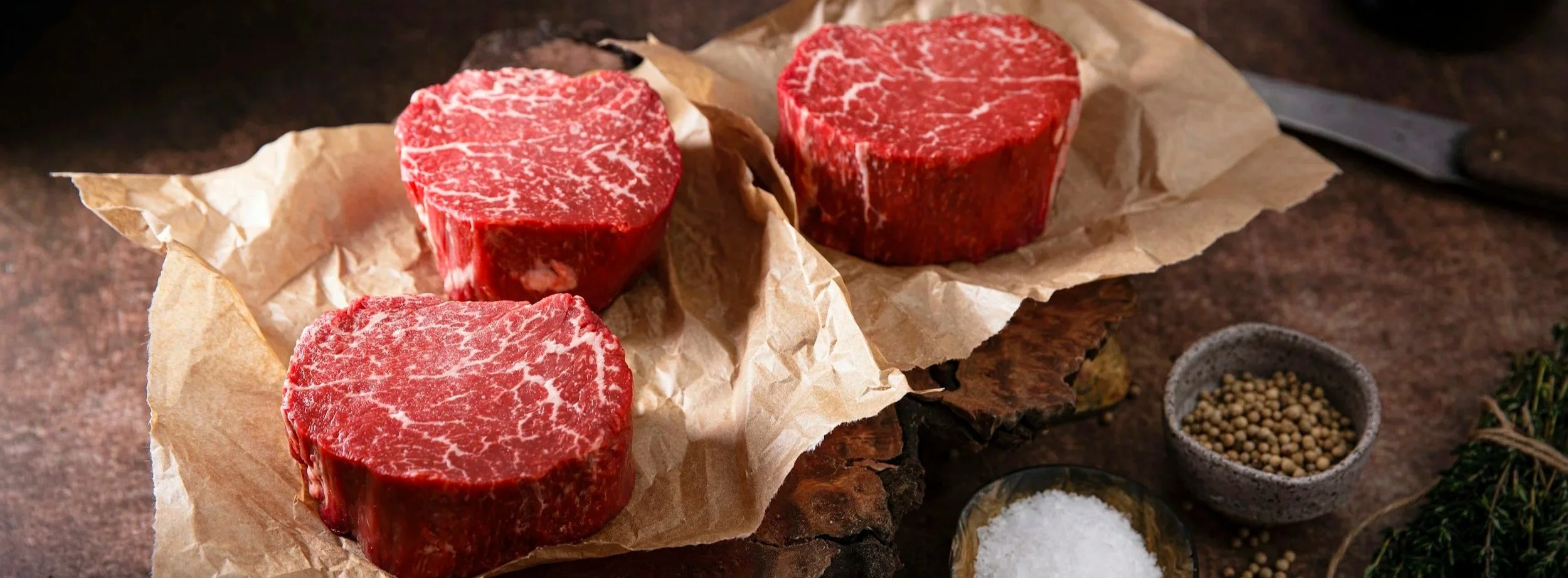 Three raw, marbled steaks on parchment paper with a knife, surrounded by bowls of salt, pepper, and herbs on a rustic wooden surface.