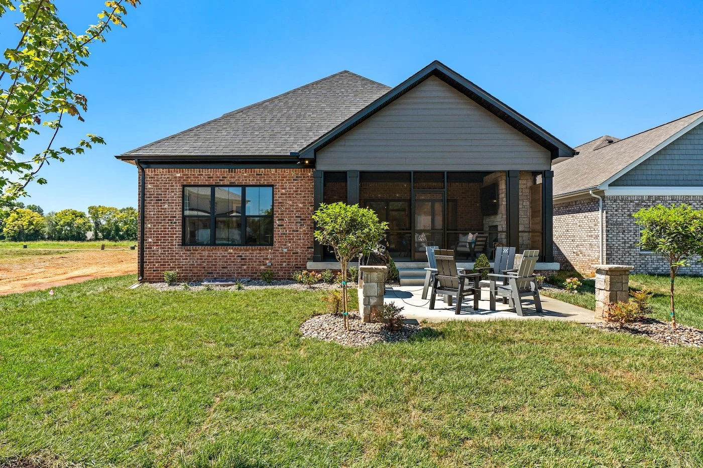 Backyard view of a house with brick and siding exterior, screened porch, outdoor dining table with chairs, green lawn, small trees, and clear blue sky.