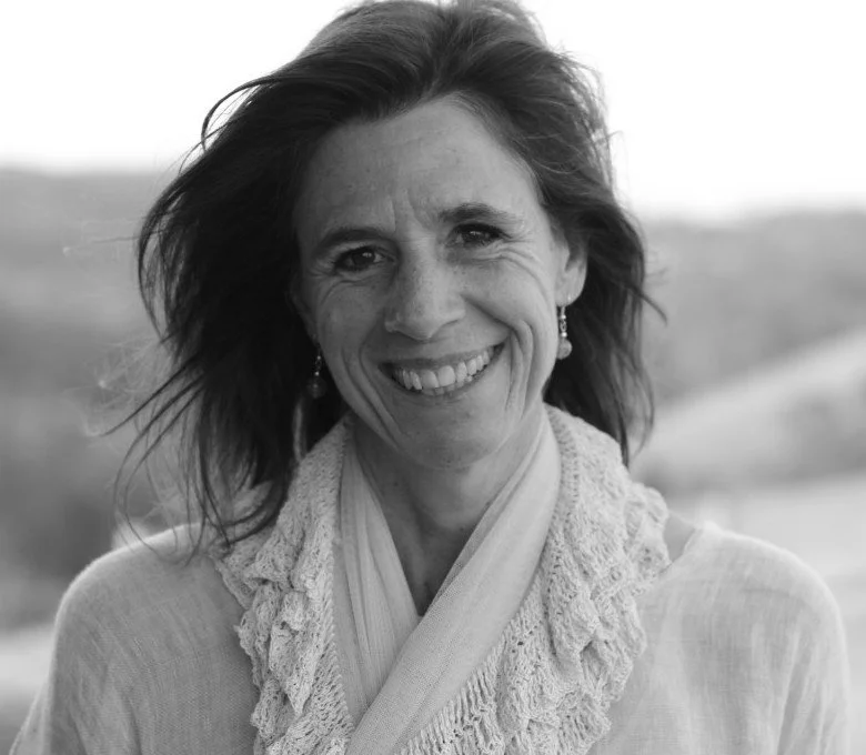 Black and white portrait of a smiling woman with shoulder-length hair, wearing earrings, a scarf, and a light-colored top, outdoors with a blurred landscape in the background.