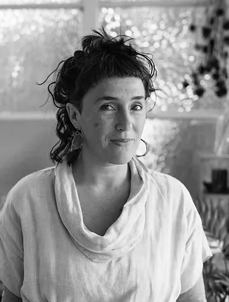Black and white photo of a woman with dark, curly hair pulled up, wearing a light-colored top, sitting indoors with a blurred background of windows and plants.