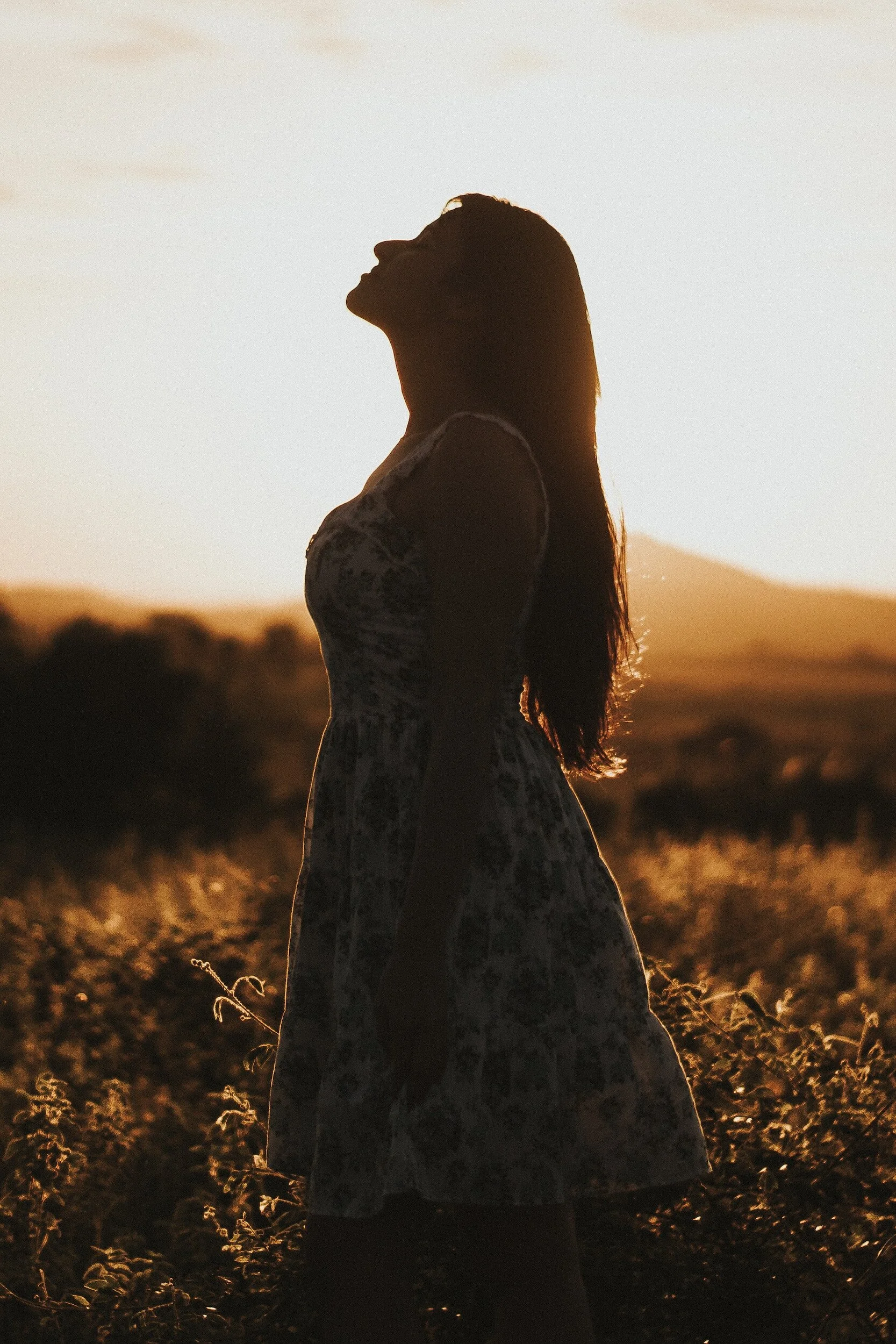 A woman standing outdoors at sunset, facing sideways with her head tilted back and eyes closed, silhouette against the bright sky.