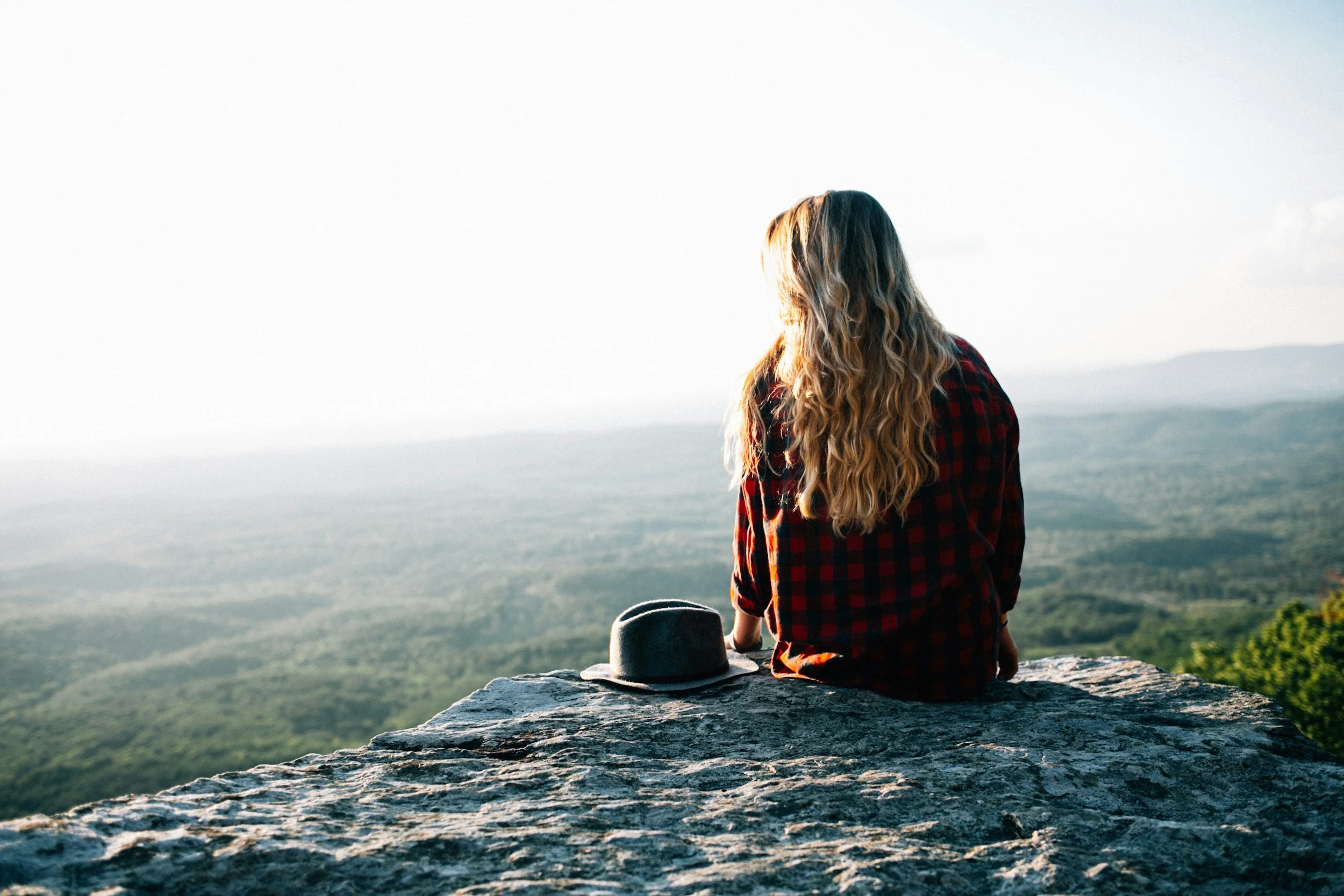 A woman with long, wavy blonde hair sits on a rocky cliff edge overlooking a vast green landscape through a hazy horizon, with a gray hat placed beside her.