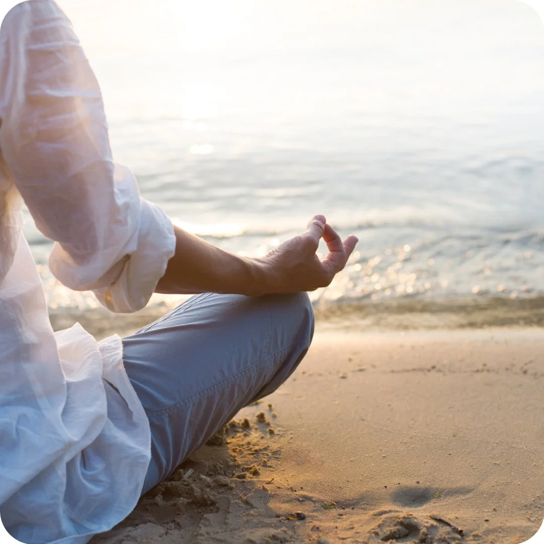 Person in white shirt sitting cross-legged on sandy beach near water, practicing meditation during sunset.