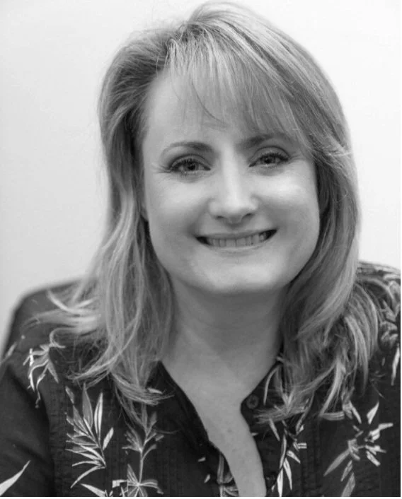 Black and white photo of a woman with shoulder-length hair smiling at the camera, wearing a patterned shirt.