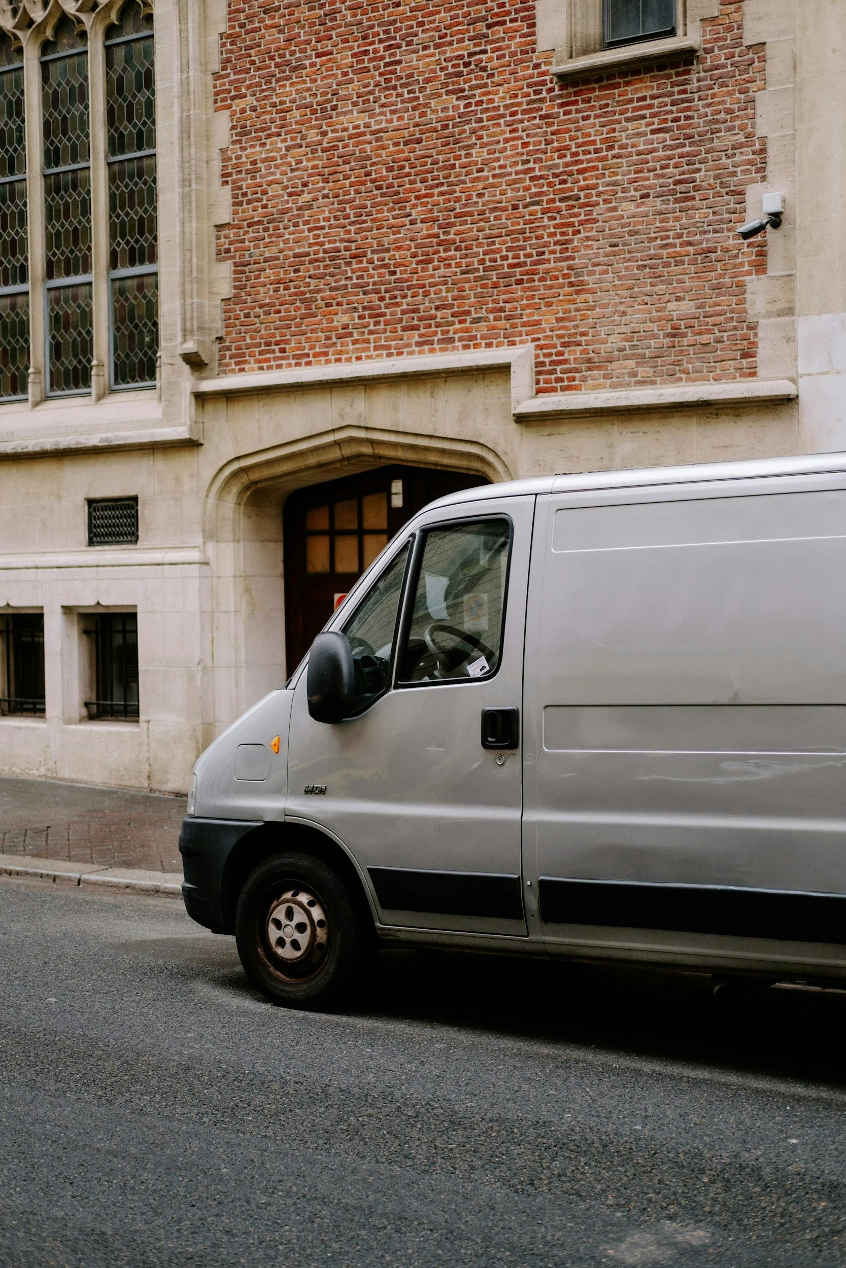 Silver delivery van parked on street in front of a brick and stone building with large stained glass windows and security camera.