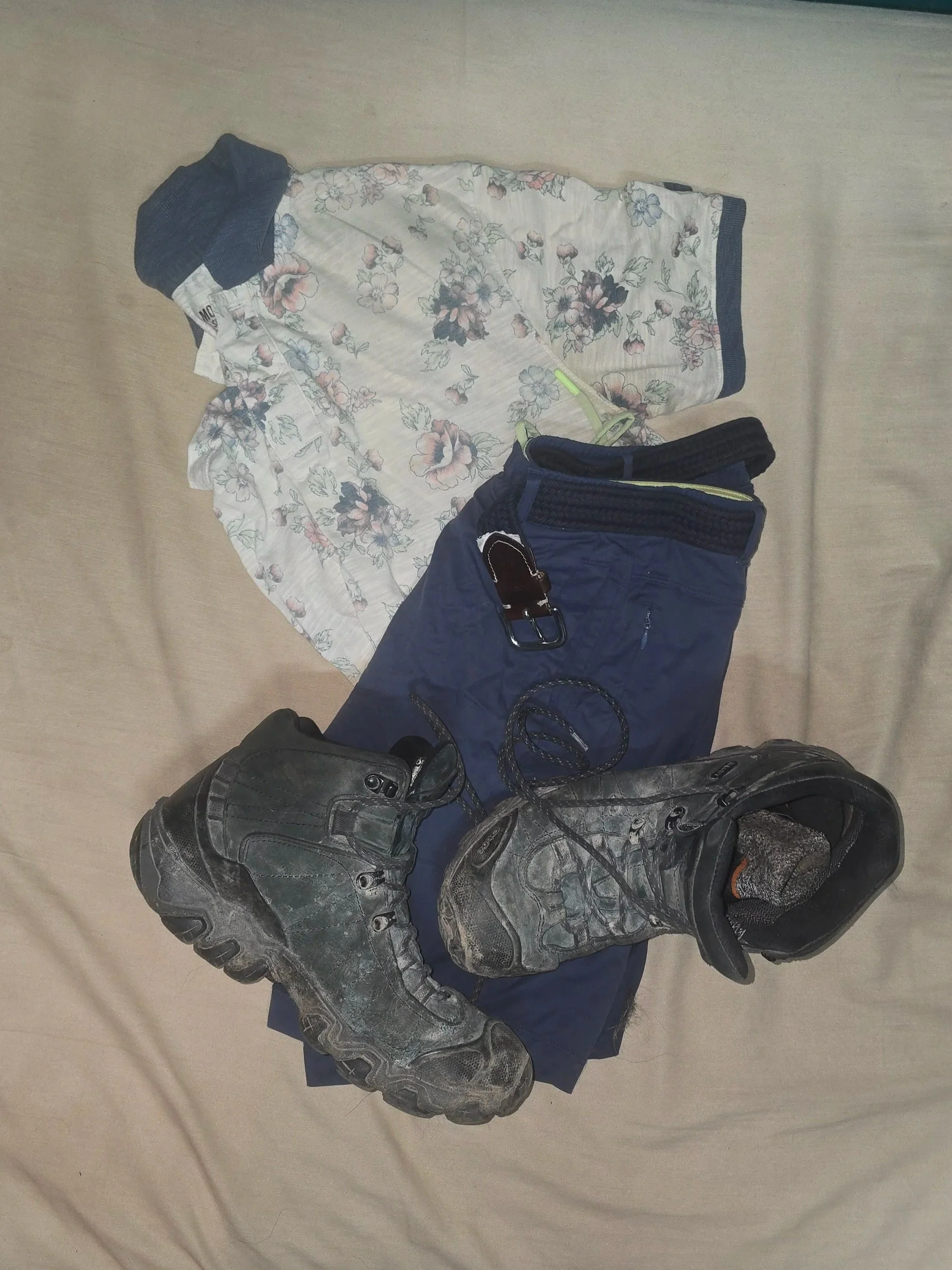 Hiking boots, blue shorts, a floral polo shirt, and a belt laid out on a bed as a casual outfit choice for a summer afternoon.