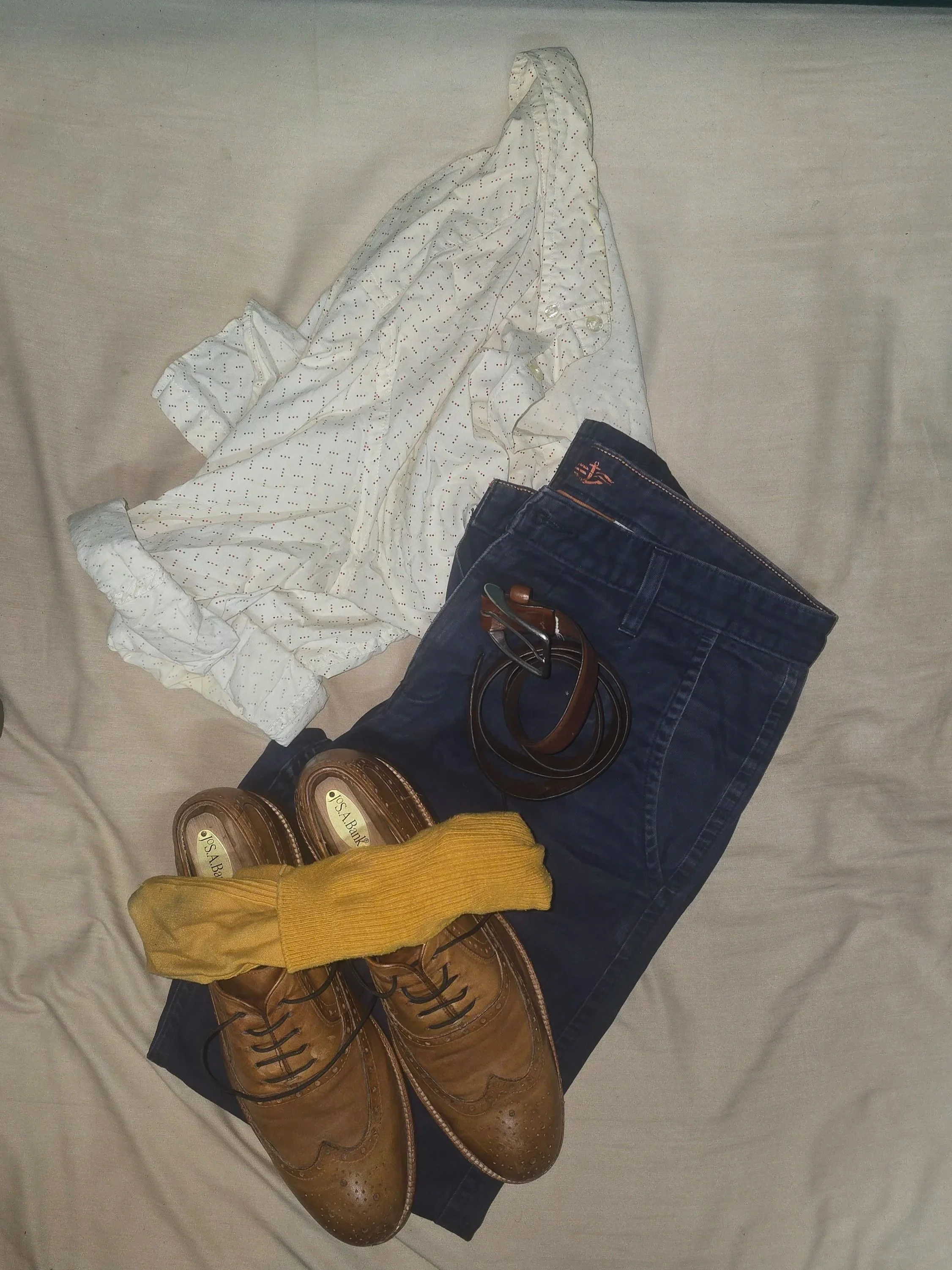 Brown brogue shoes, navy chinos, yellow socks, and a patterned button-down shirt arranged on a bed as a colorful business-casual outfit.