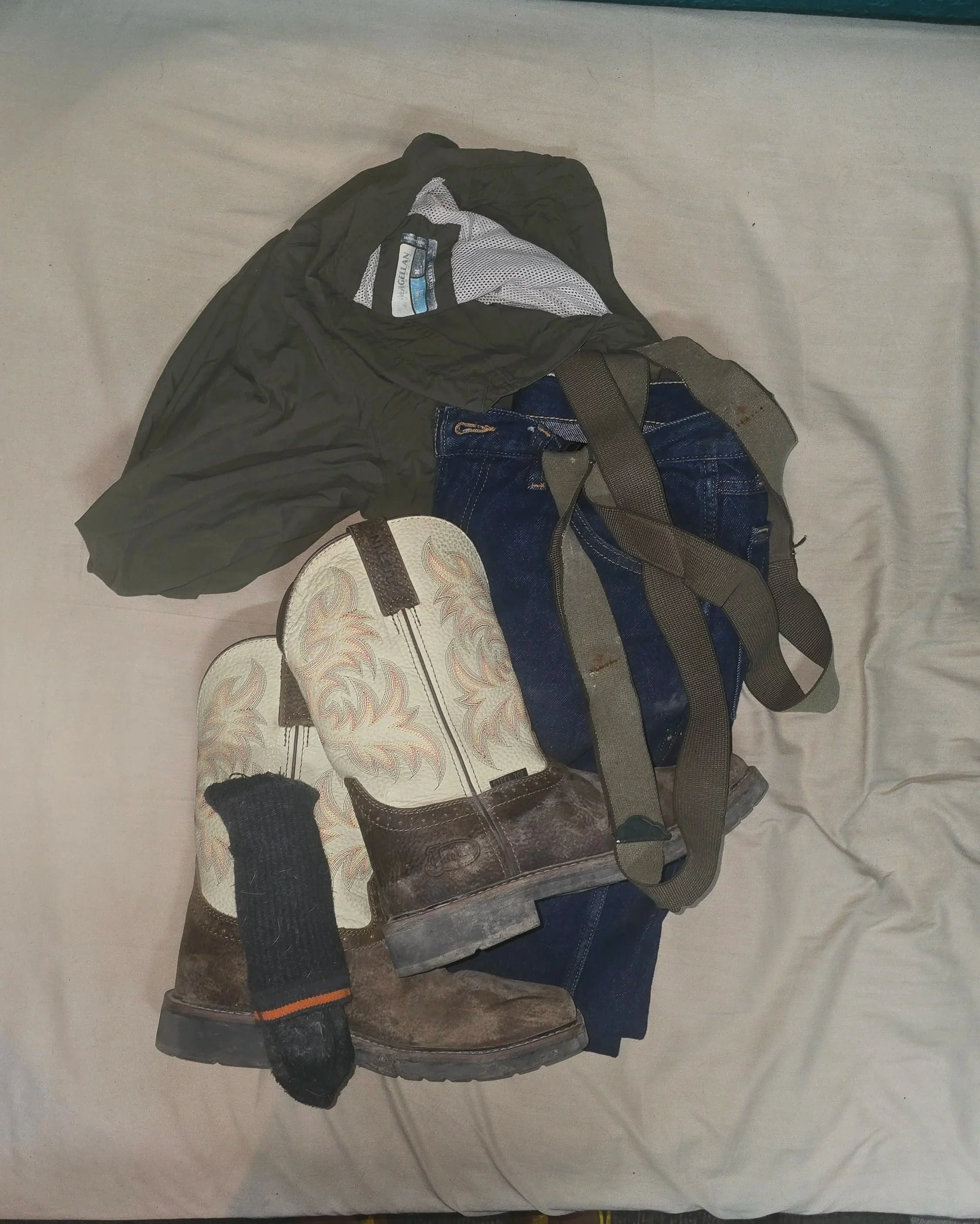 Cowboy boots, jeans, suspenders, and a fishing shirt arranged on a bed to form a country workwear outfit.