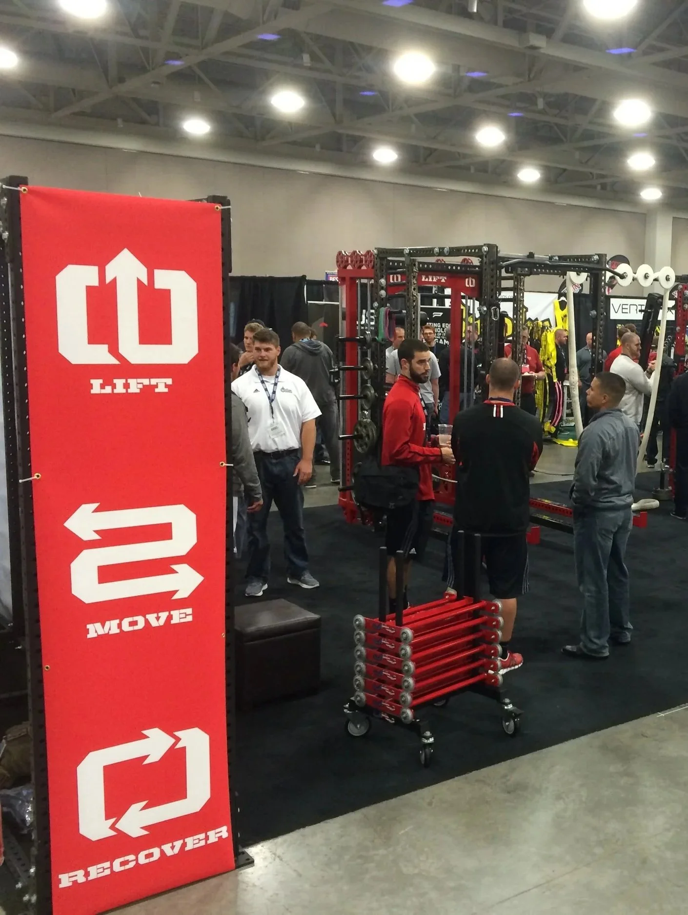 Fitness equipment display at a trade show with a red vertical sign showing Lift, Move, and Recover directions, with several people interacting and examining fitness gear and racks in the background.