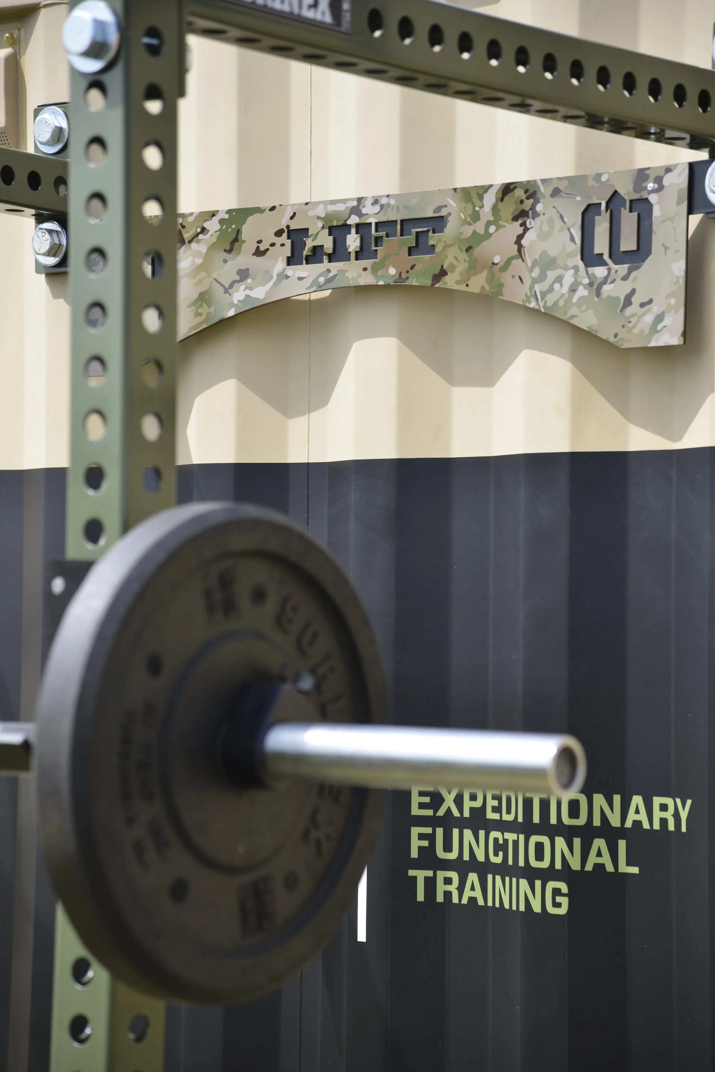 Close-up of a weightlifting barbell with a weight plate attached, set up in front of a squat rack logo arch that reads 'MOVE' with a camo pattern