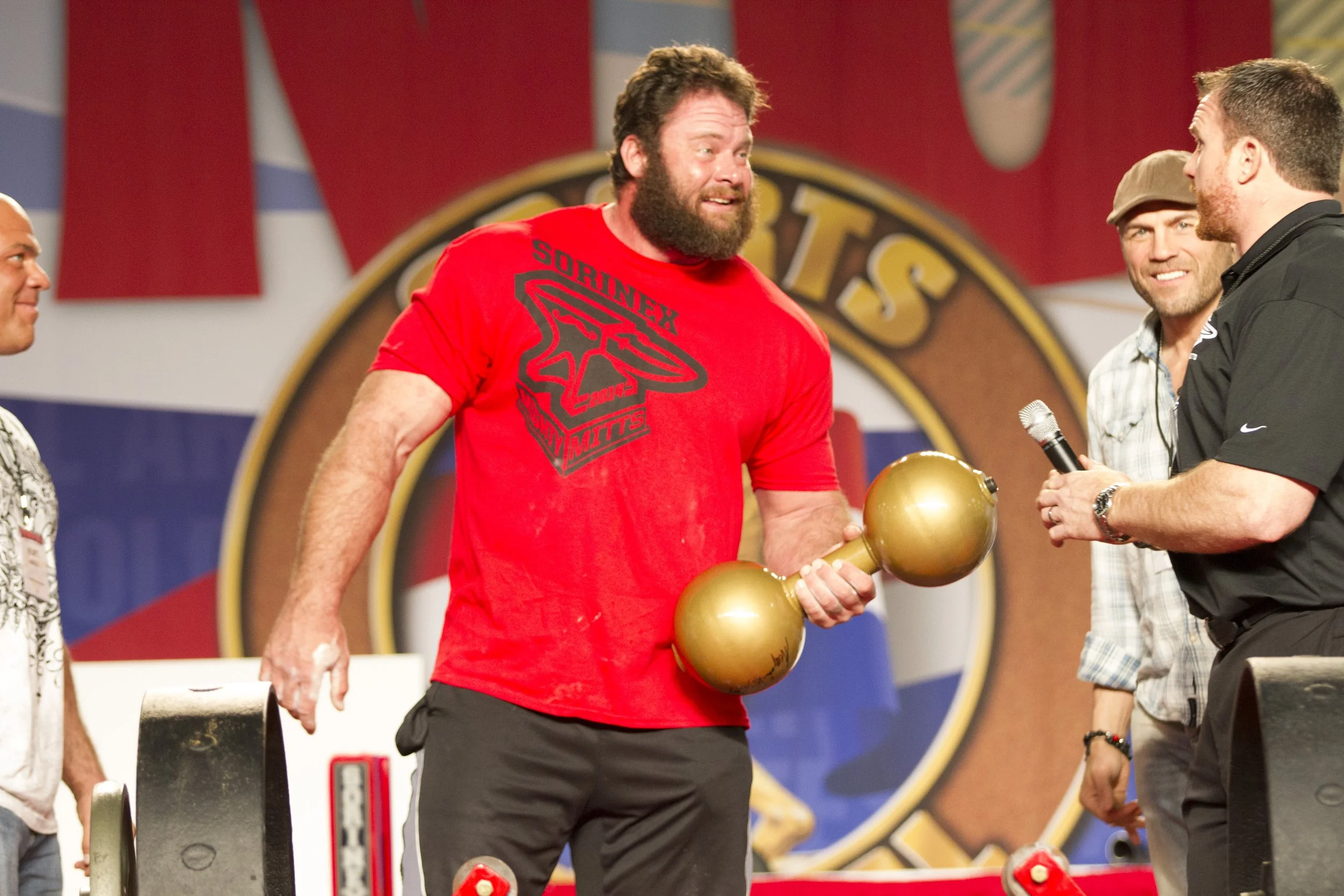 A muscular man with a beard wearing a red Mighty Mitts T-shirt holding a gold dumbbell on stage at the Arnold Strongman competition.