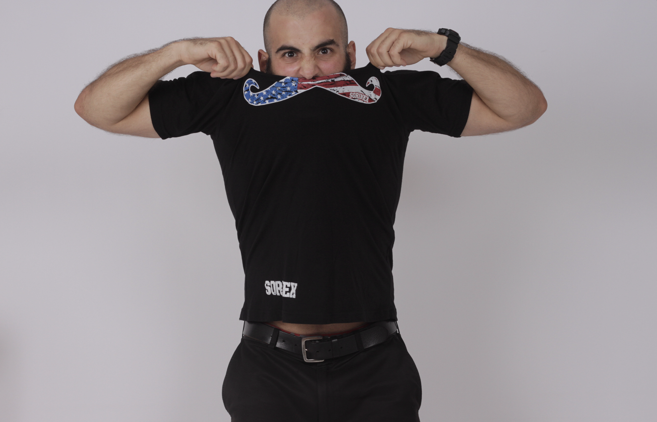 Man with a shaved head lifting his black T-shirt to show the american flag moustache logo over his face while smiling