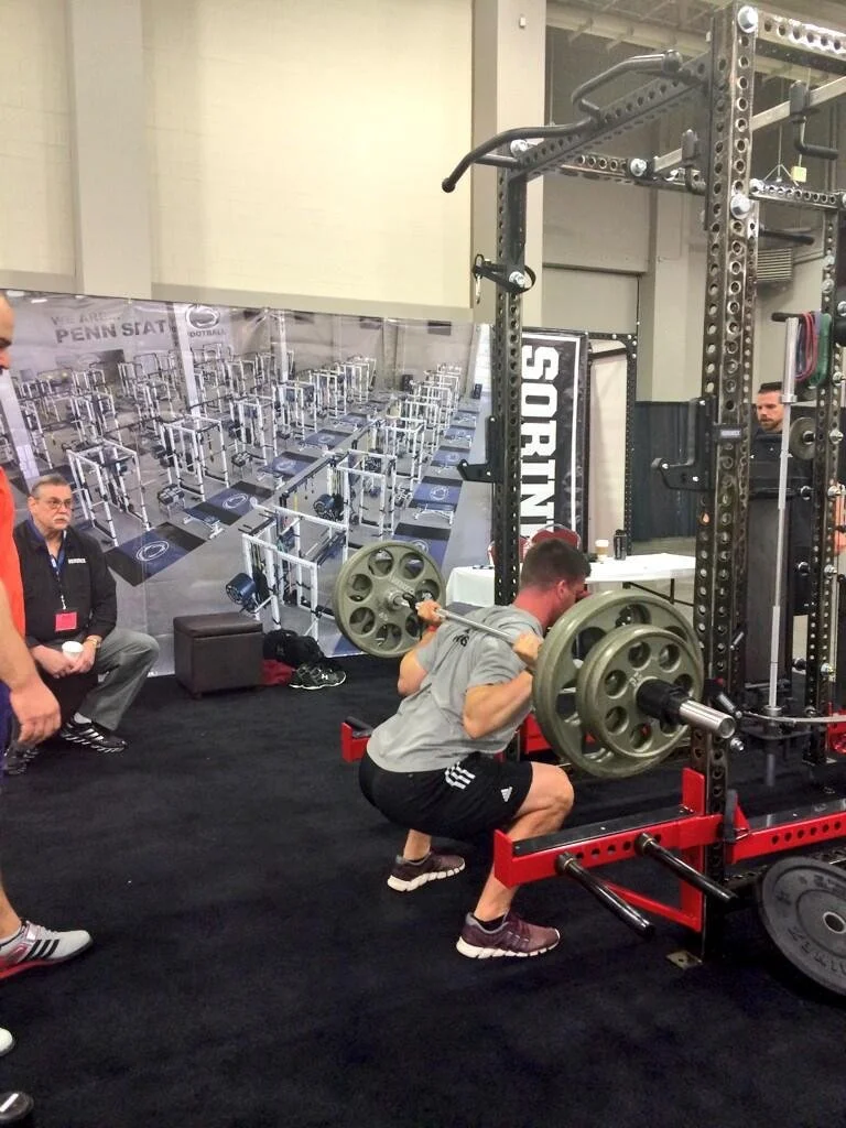 Man performing a barbell squat in a gym or fitness expo, with people watching.