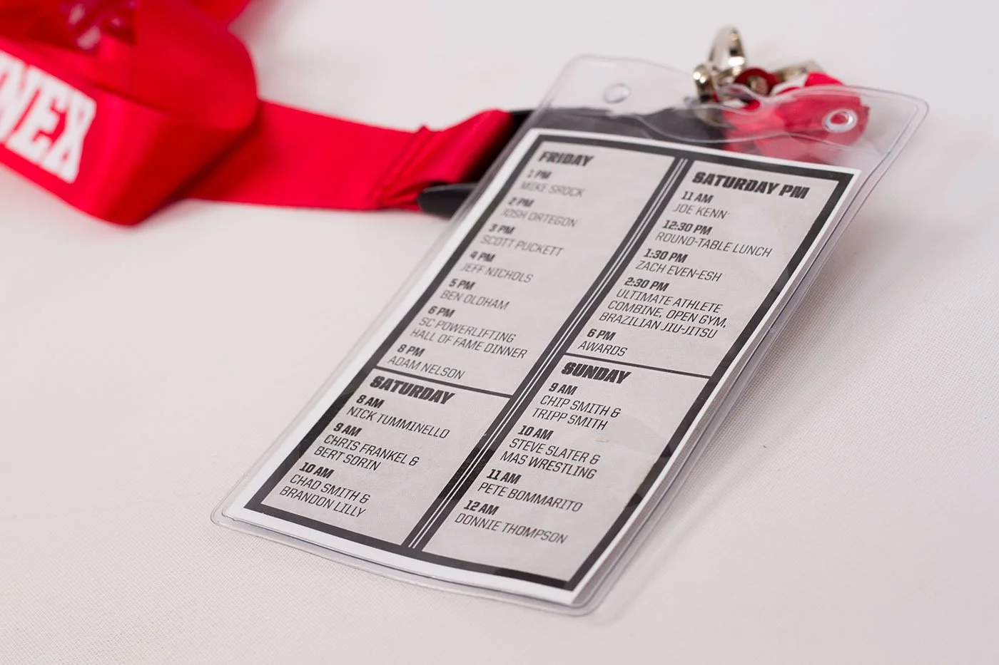 Detailed view of back of Summer Strong name badge showing event schedule with times and names, enclosed in a clear plastic holder, alongside a red lanyard with a badge at a white surface.