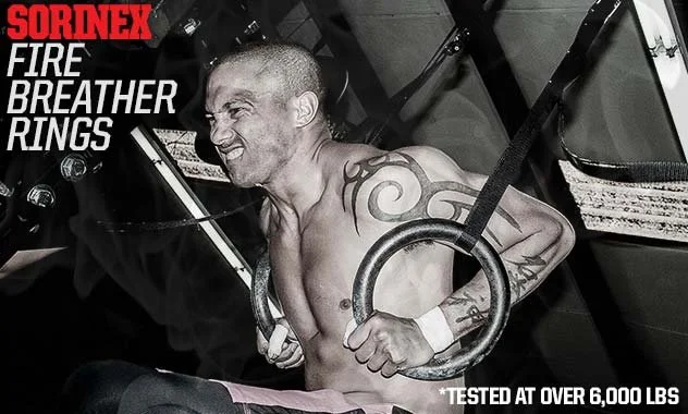 A shirtless man with tattoos on his shoulder and arm flexes muscles while holding gymnastic rings during a workout or fitness event. The image is in black and white with red and white text that reads 'SORINEX FIRE BREATHER RINGS' and smaller text stating '*TESTED AT OVER 6,000 LBS'.