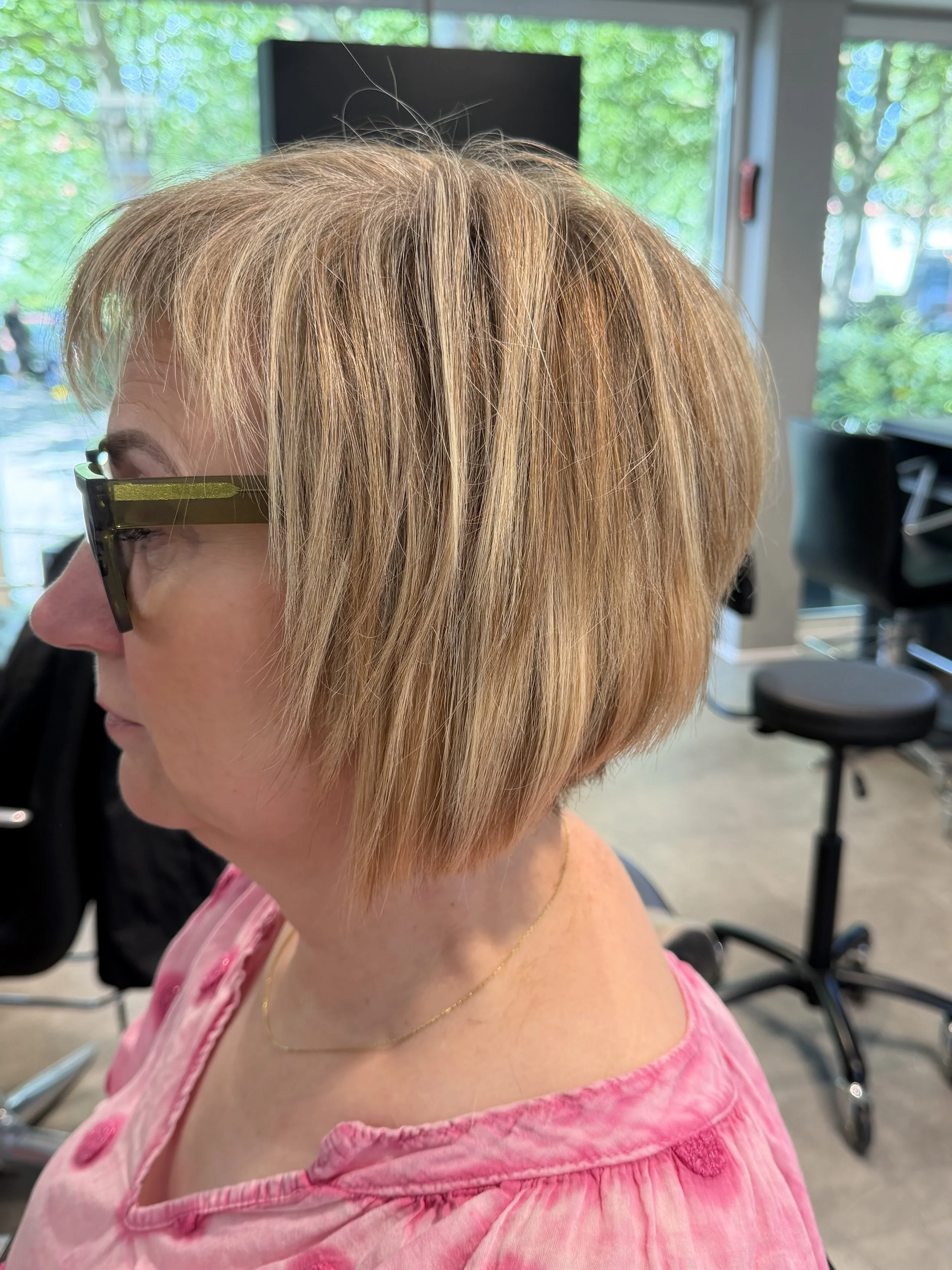 Vi laver mange extensionsbehandlinger til kvinder med kort hår og page, da det giver mere fylde og volume. Et kvindes hoved med kort, blond, let rodet hår, set fra siden, i et frisørsalon med store vinduer og udstyr i baggrunden.
