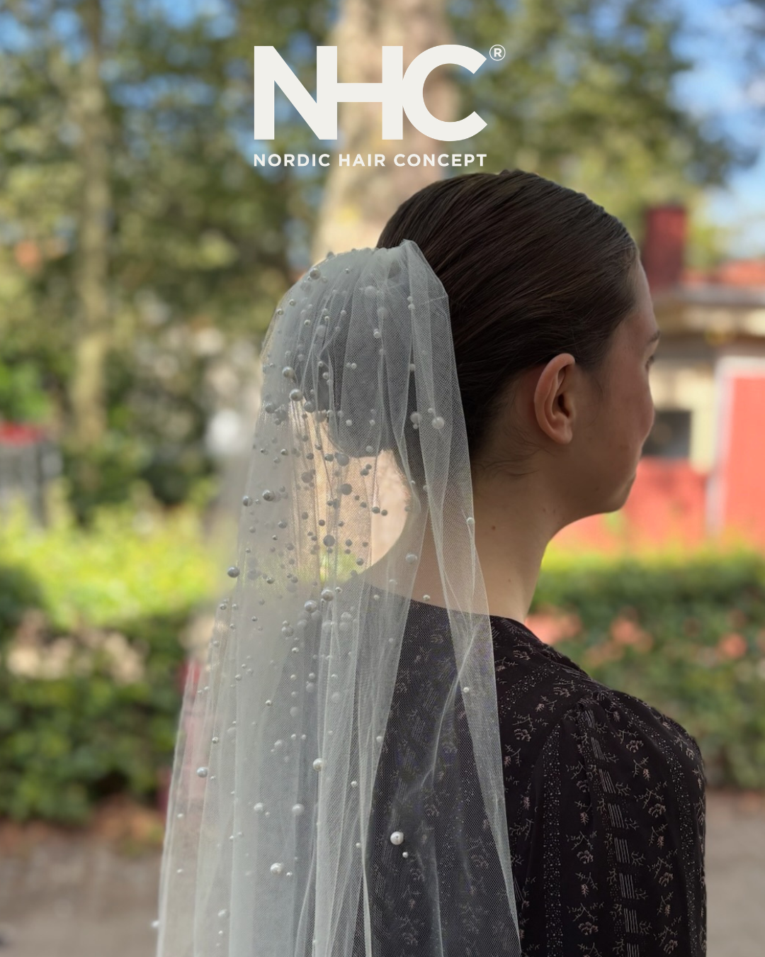 Profil af en kvinde med glat, mørkt hår iført en brudekjole og en tyllet veil med perler, mod en baggrund af træer og bygninger.