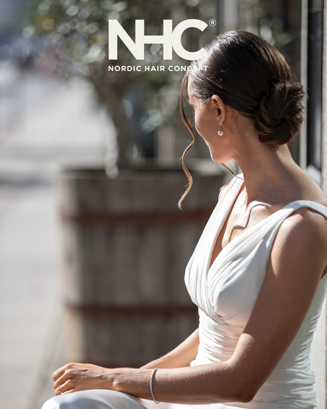 En kvinde i en hvid kjole sidder udenfor med hendes ryg til kameraet. Hendes hår er opsat med en løs krøllet fnug i blødt sollys. Baggrund med udeplanter og cafébord.