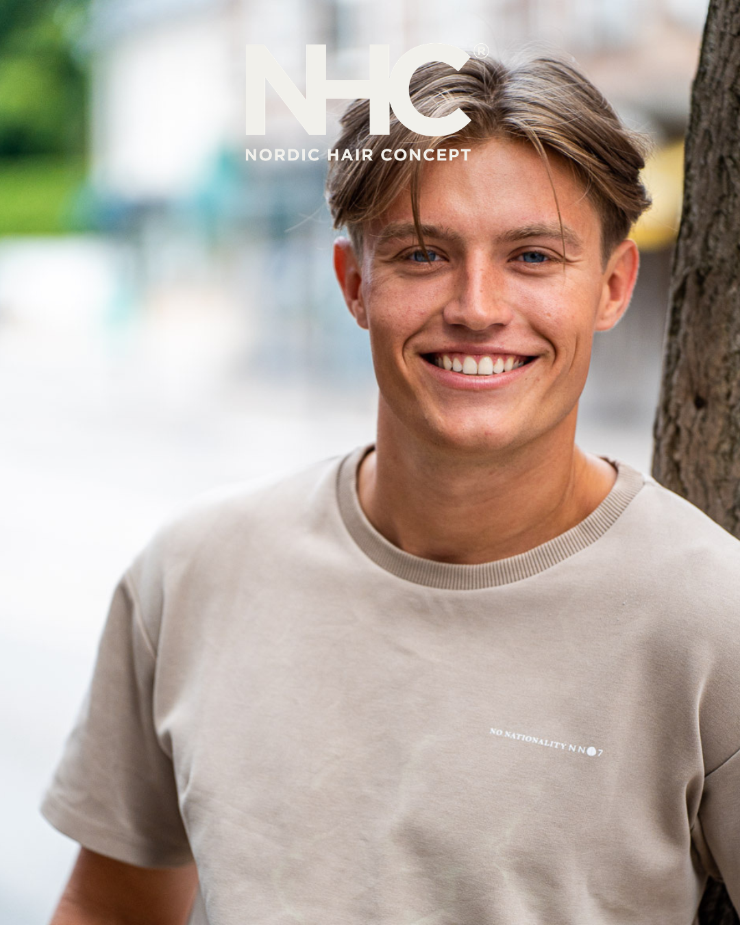 Ung mand smiler foran en træstamme, iført en beige T-shirt med teksten 'No Nationality No 7'.