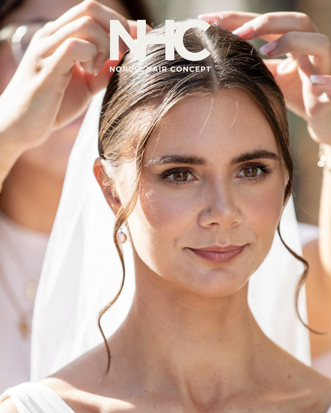 En smuk brud med brunt hår får hjælp til sin bryllupshår fra en person med hænder i baggrunden.